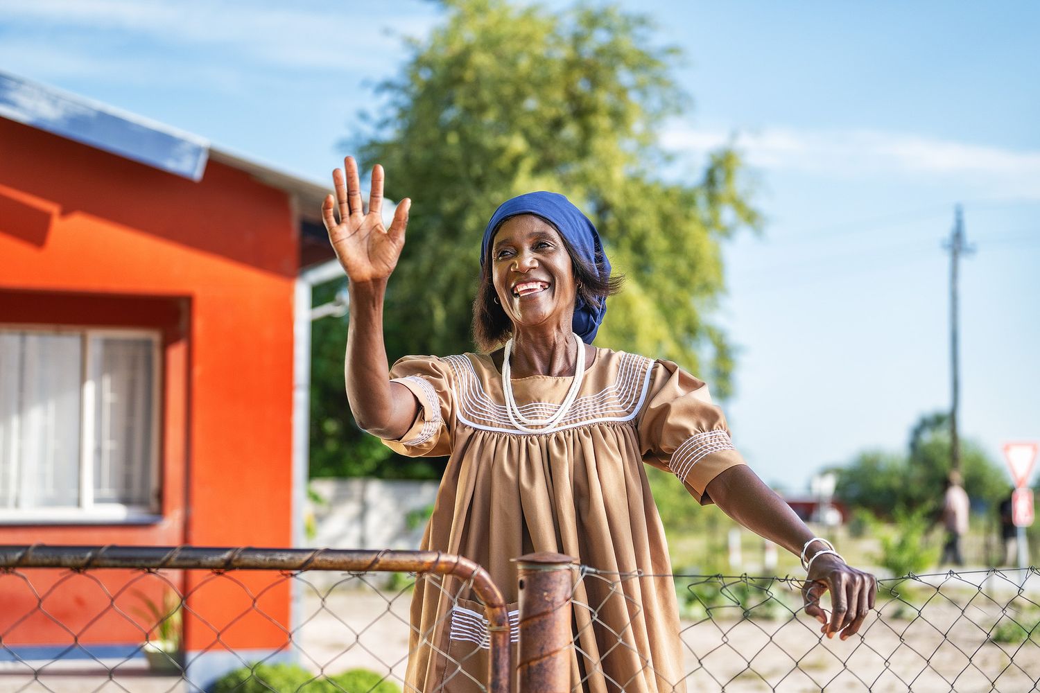 A smiling woman in traditional African attire waves cheerfully near a fence outdoors.