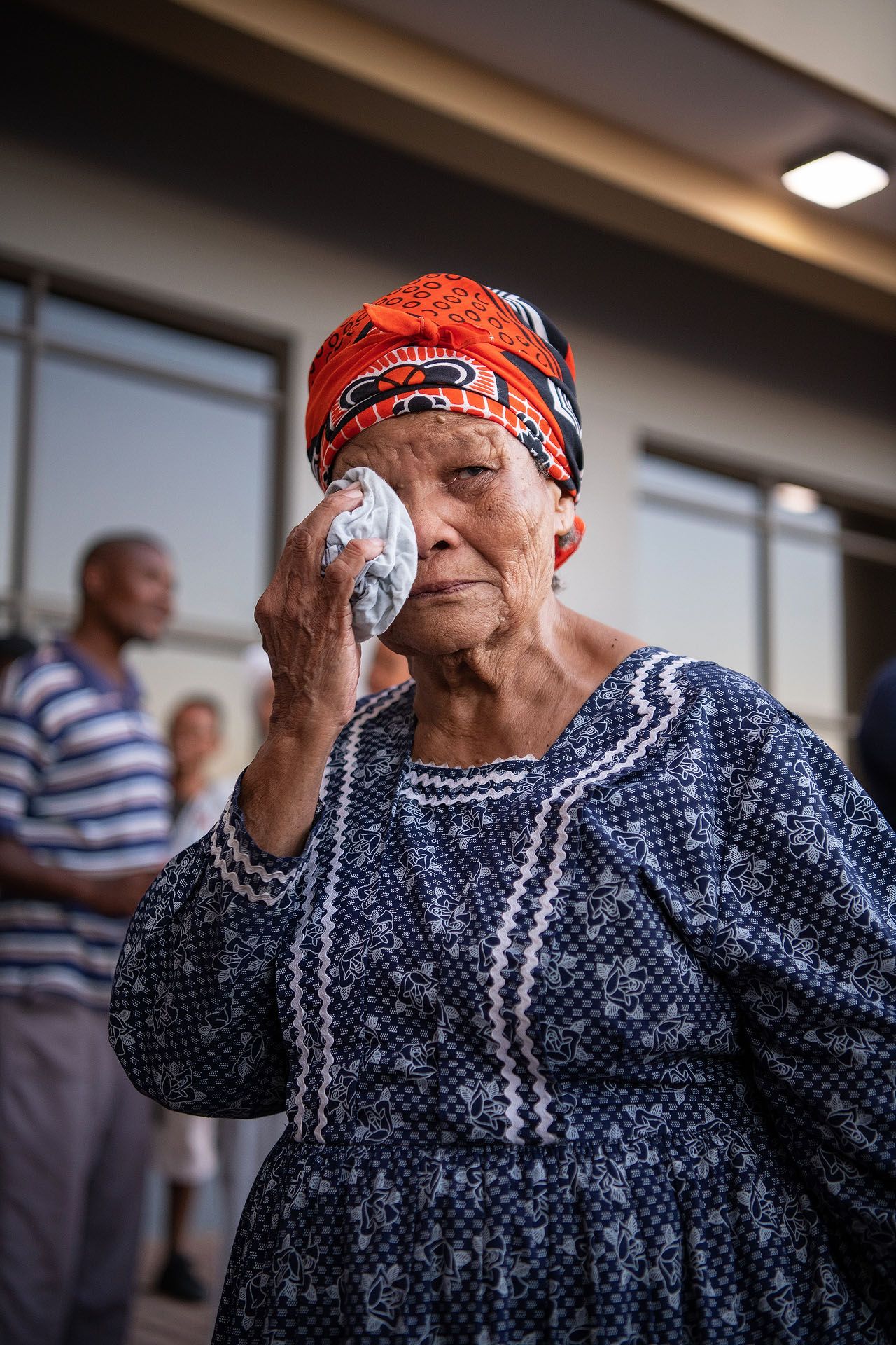 Elderly woman in blue dress and red headscarf holds cloth to her eye outdoors.