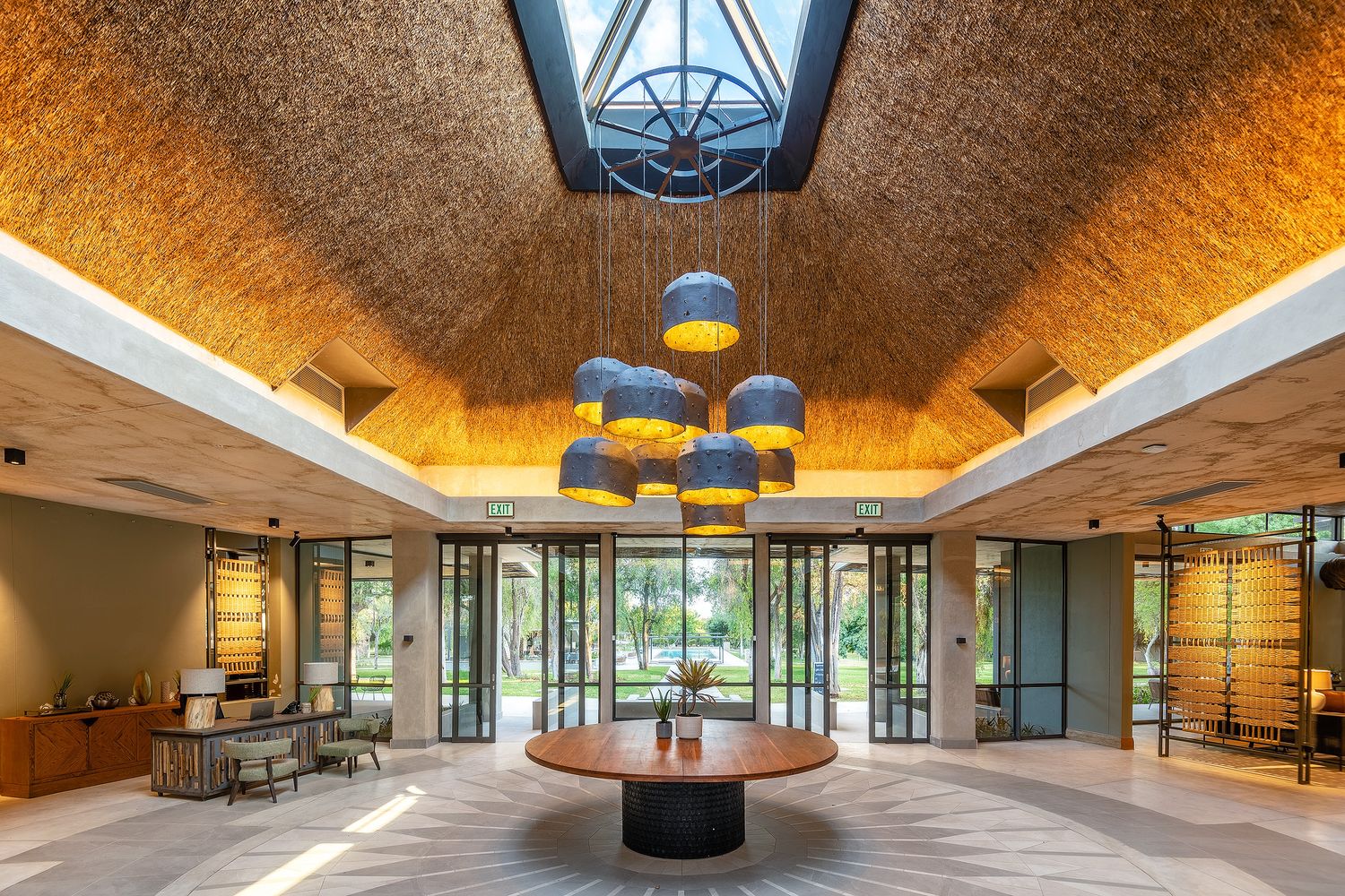 Elegant hotel lobby with textured gold ceiling, blue pendant lights, round table, and glass doors leading to lush greenery.