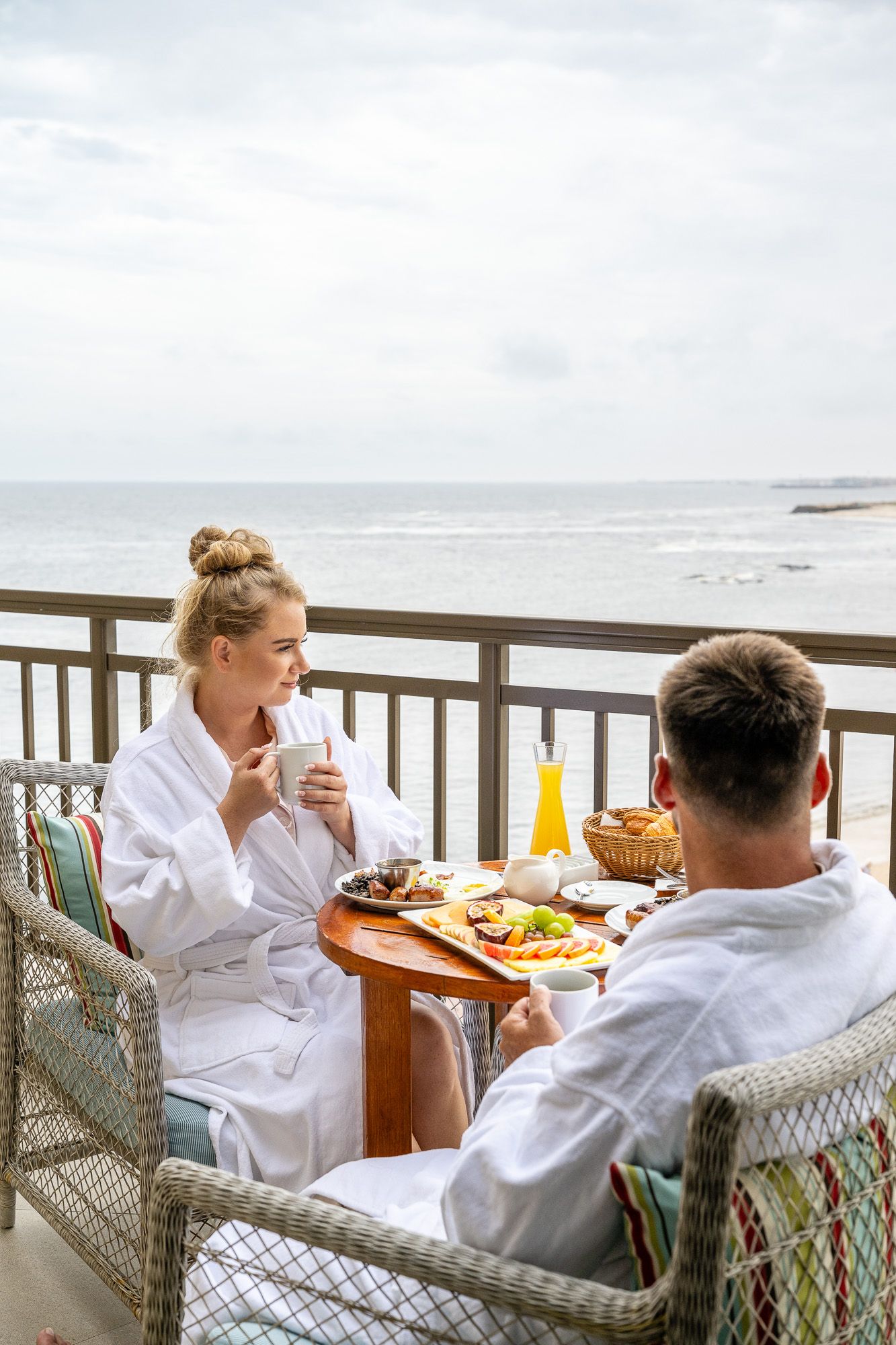 Couple in white robes enjoying breakfast on a beachfront balcony with ocean views and fresh juice.
