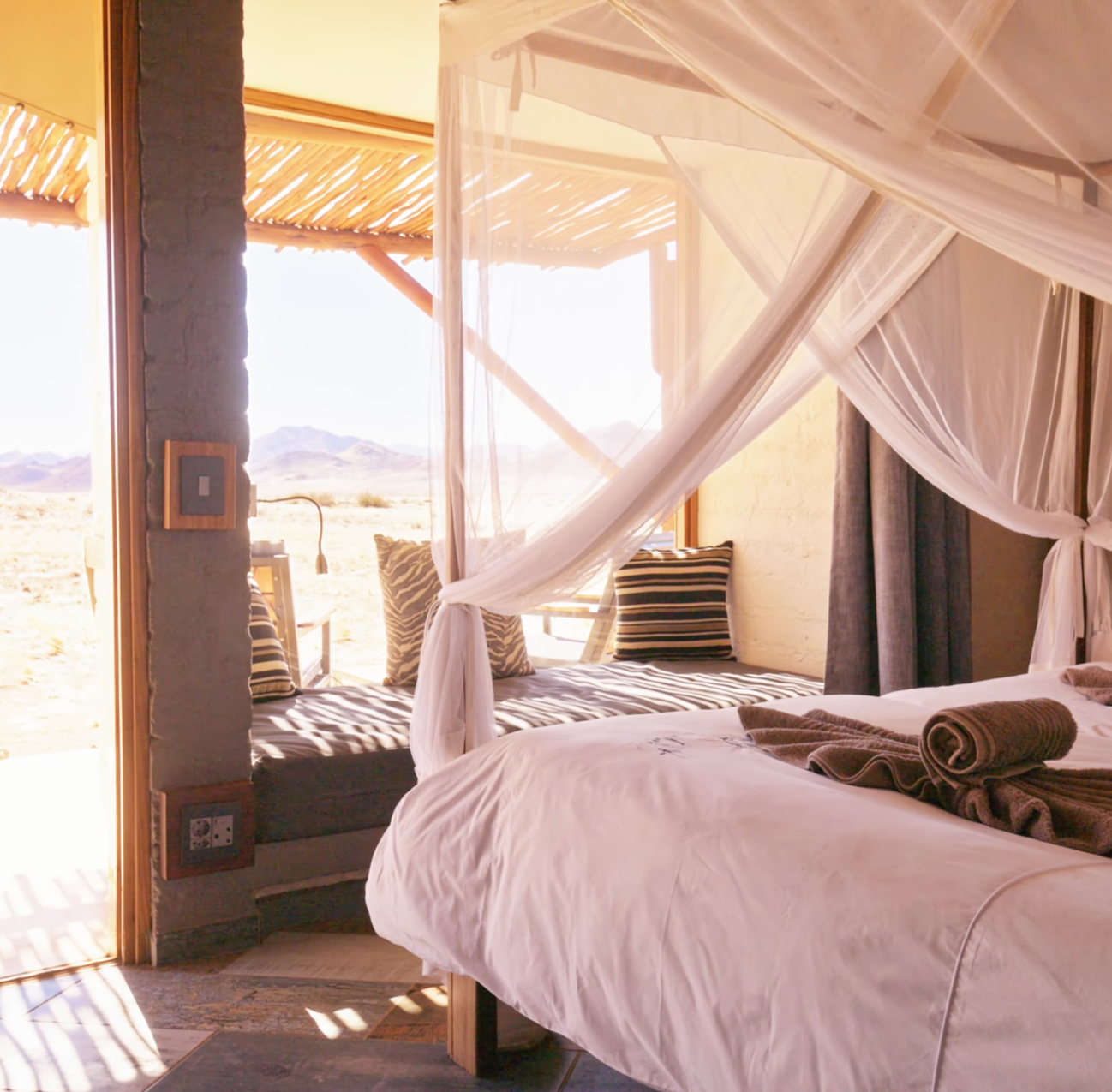 Luxury safari bedroom with white canopy bed, striped pillows, and open doors revealing a stunning desert landscape view.