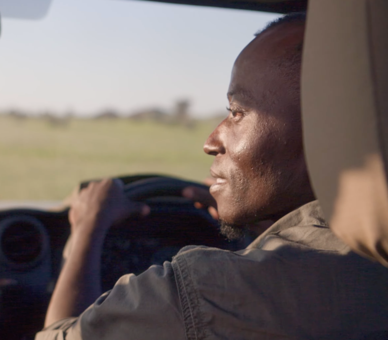 A man in a khaki shirt drives a safari vehicle across an open savanna landscape.