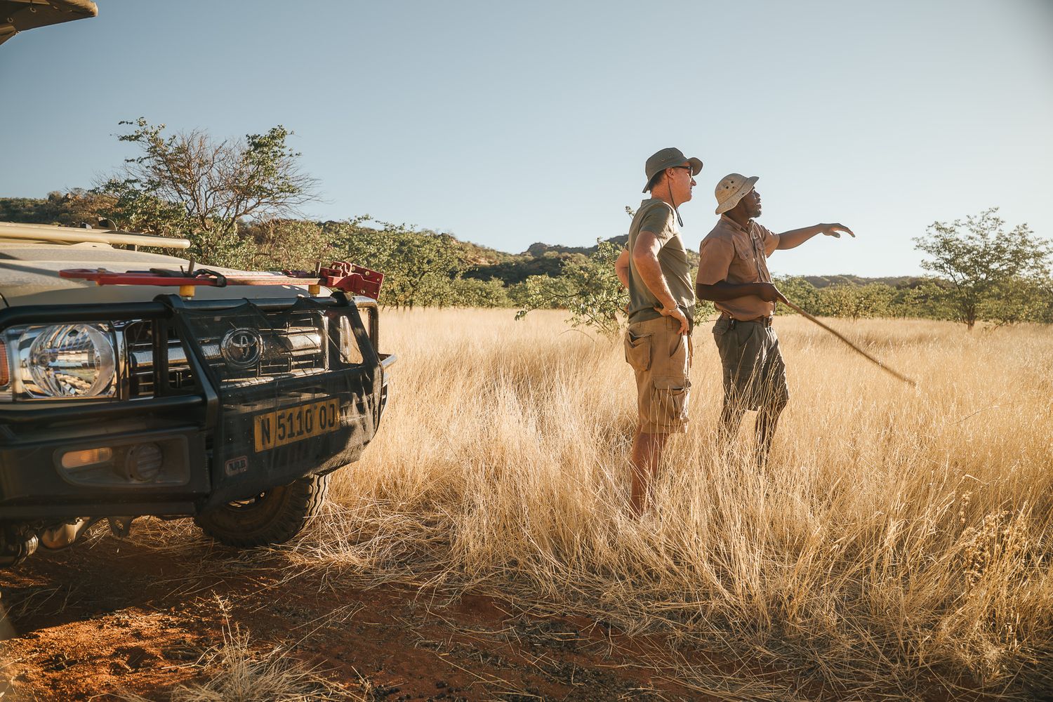 Two safari guides stand beside a jeep in golden grasslands, one pointing into the distance on a sunny day.