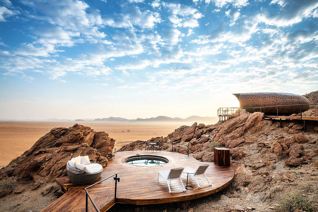 Elevated rocky desert retreat with circular hot tub, wooden deck, and sweeping arid landscape views at sunrise.
