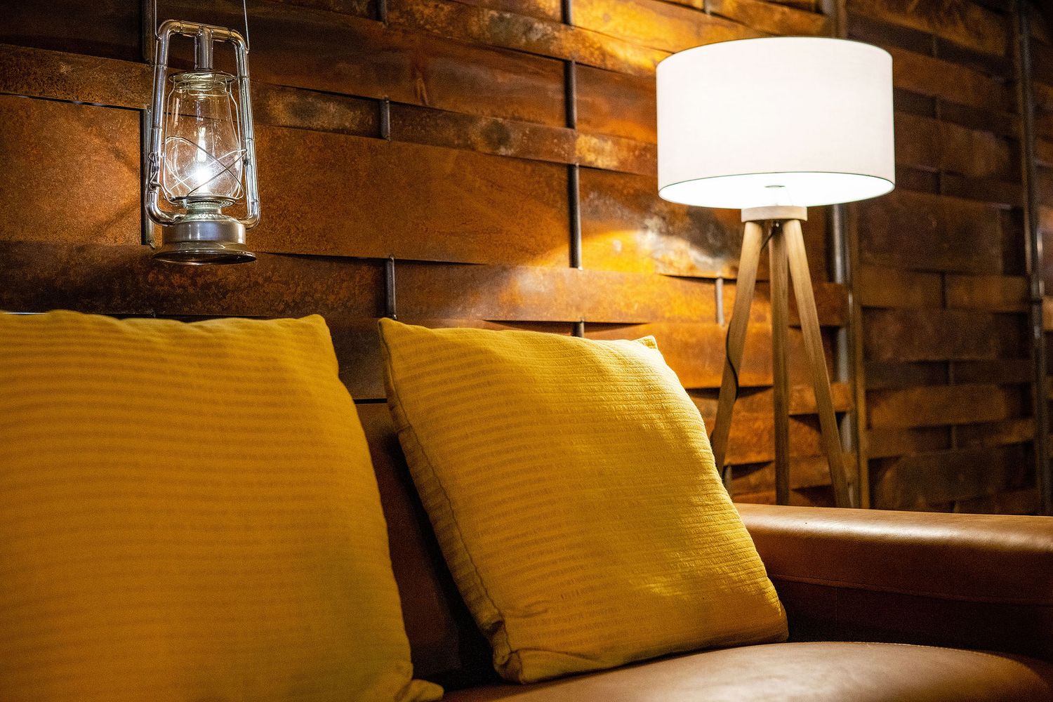 Cozy living room corner with yellow pillows on leather sofa, wooden tripod floor lamp, and rustic brick wall with lantern.