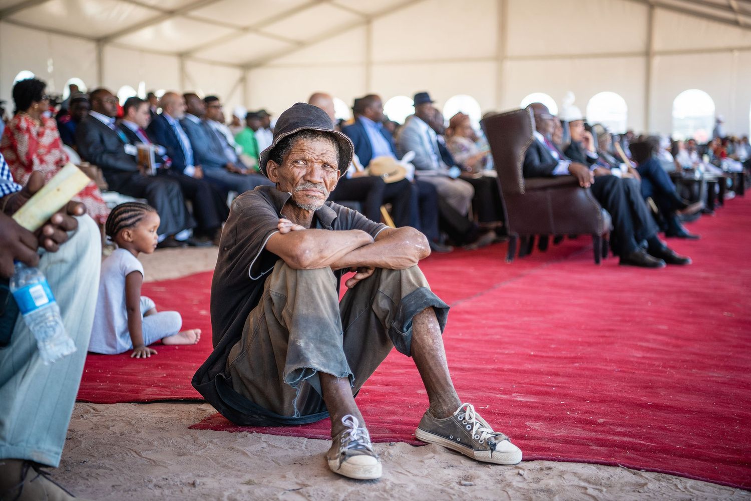 A man in a hat sits cross-legged on a red carpet at a large outdoor gathering under a white tent.