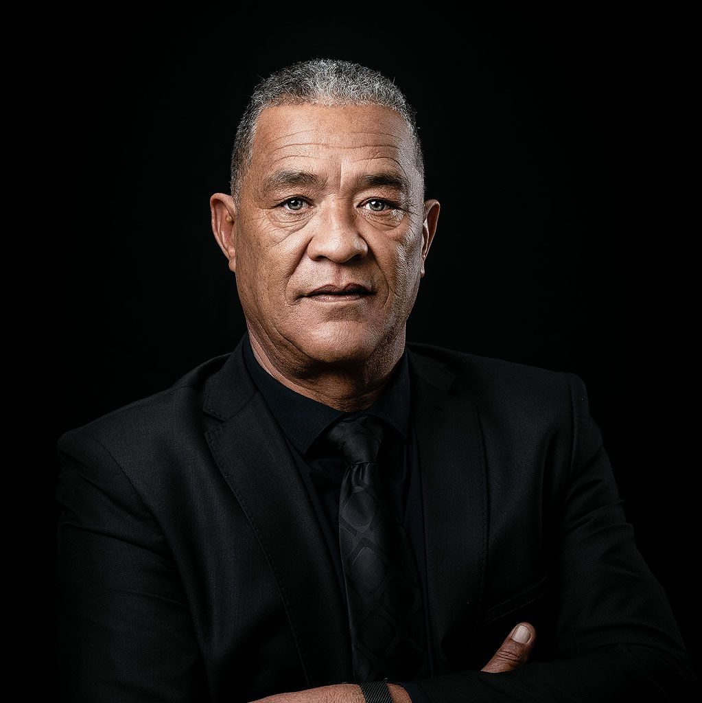 A confident middle-aged man in a black suit and tie poses with arms crossed against a dark background.