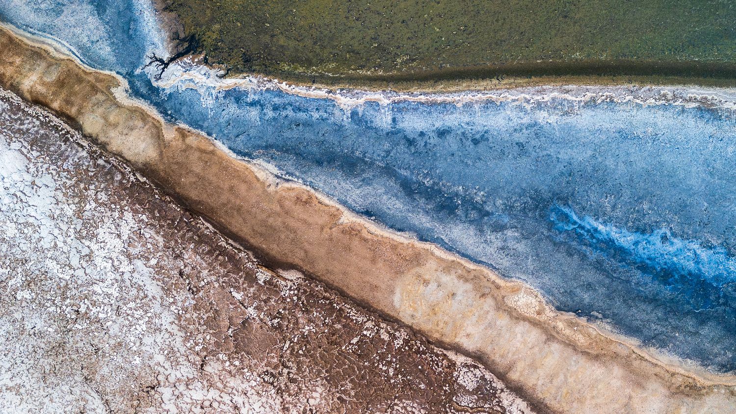 Aerial view of a salt lake shoreline with striking blue water, white salt deposits, and brown earth creating vivid natural patterns.