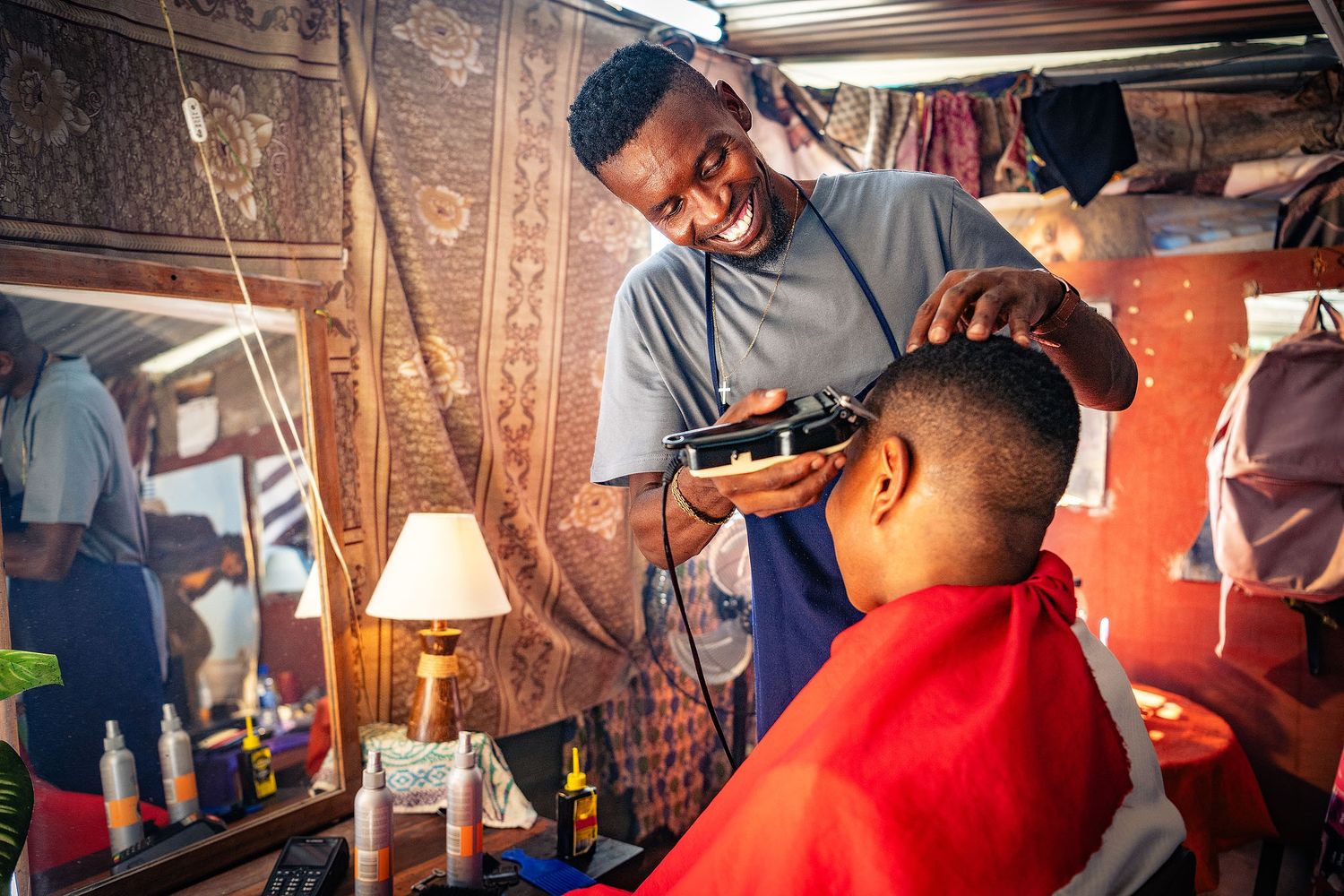 A smiling barber uses clippers to cut a young boy's hair in a colorful barbershop with mirrors and products.