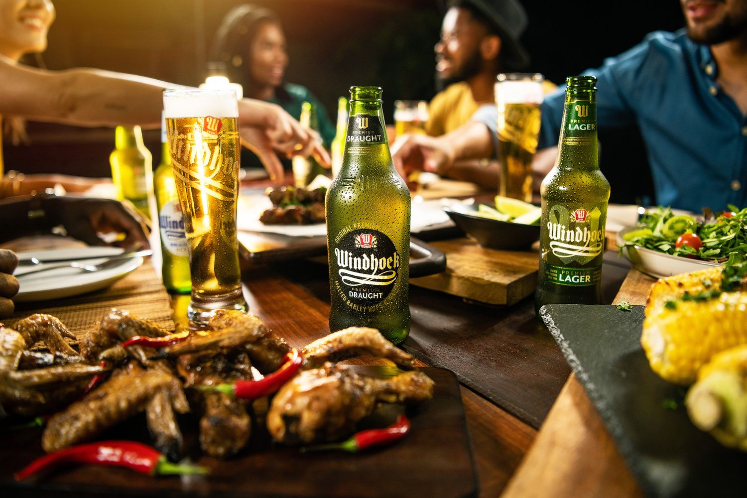Friends dining together with Windhoek Lager beer bottles and glasses on a table full of grilled food.