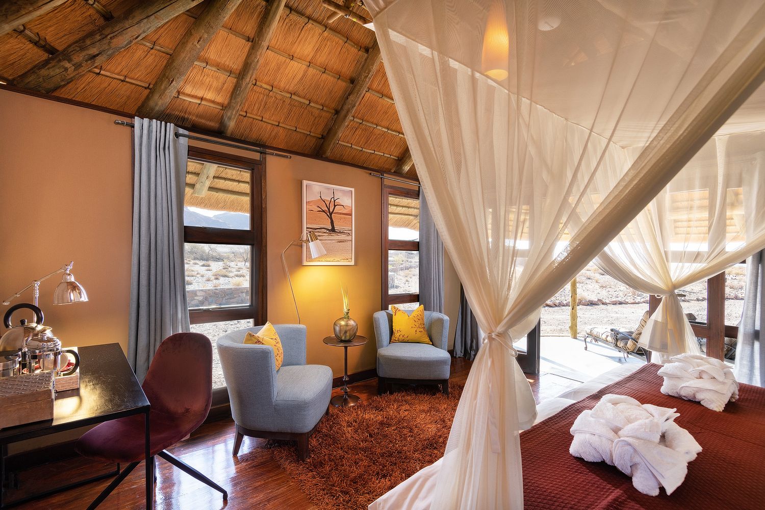 Luxury safari lodge room with canopy bed, blue armchairs, wooden floors, and thatched ceiling overlooking African landscape.