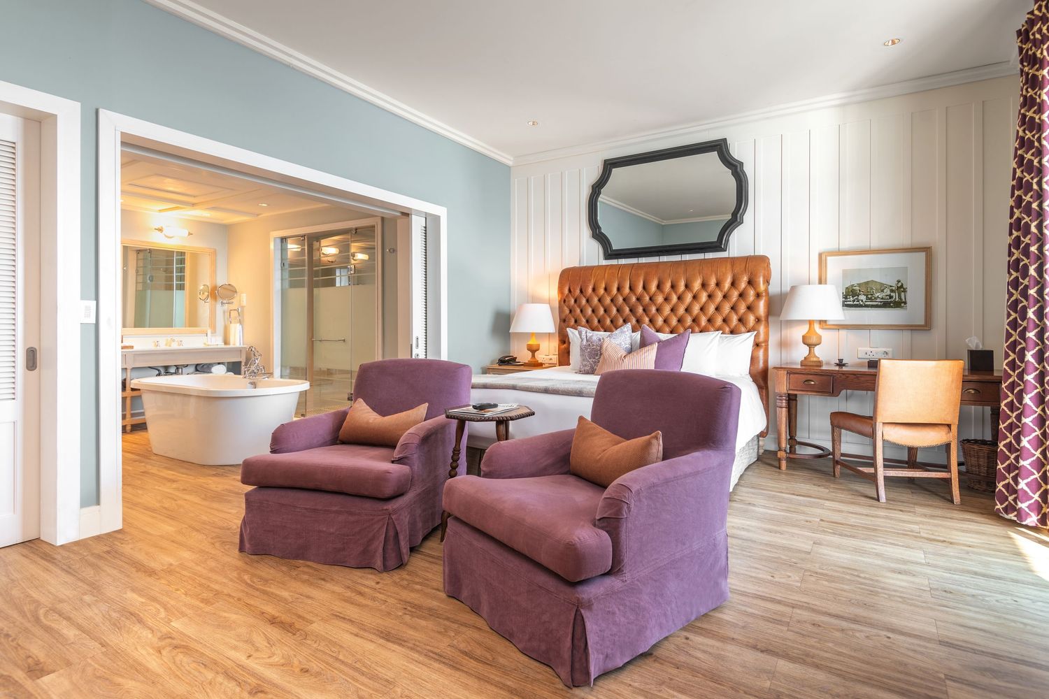 Elegant hotel suite with tufted orange headboard, purple armchairs, hardwood floors, and en-suite bathroom with freestanding tub.