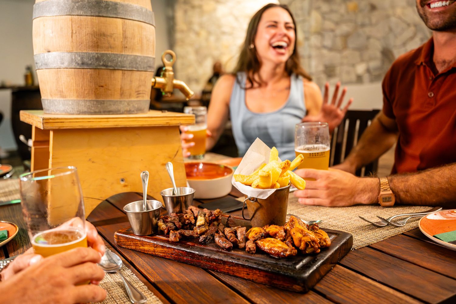 Friends laughing and sharing grilled meat platter with chips and cold beers at a rustic restaurant table.