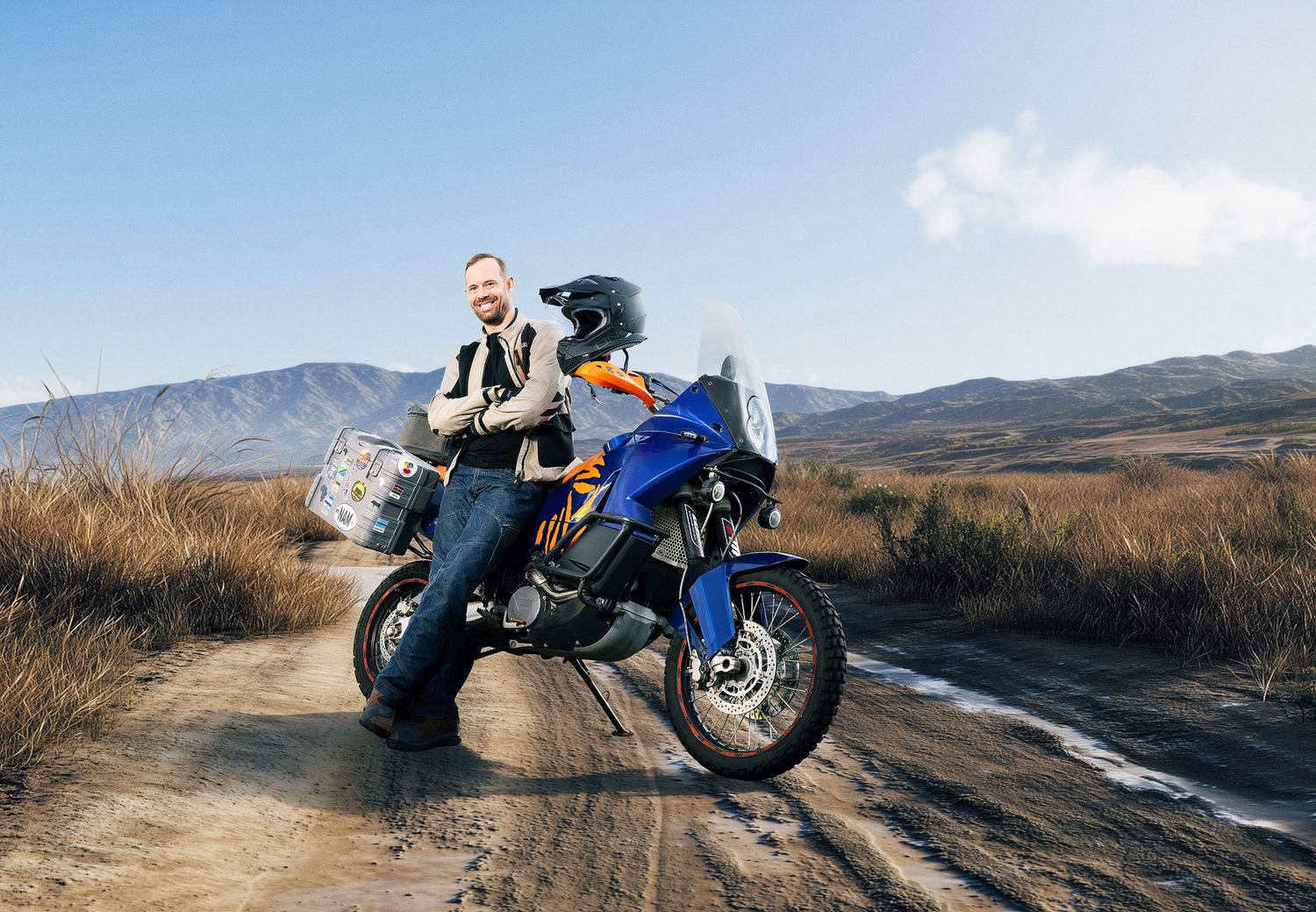 Man posing with a blue adventure motorcycle on a dirt road with mountain landscape in the background.