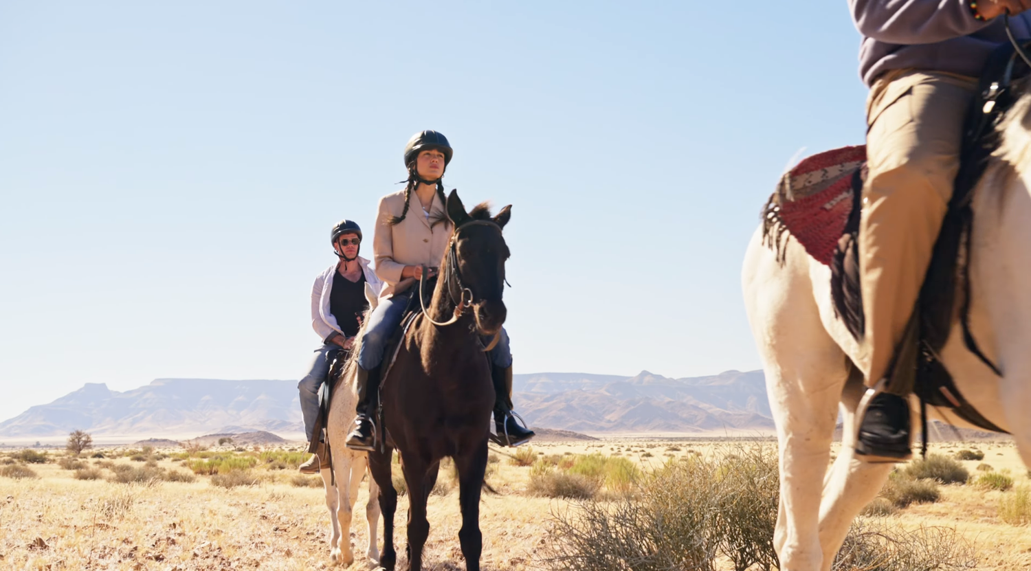 Group of riders on horseback trekking through a desert landscape with mountains in the background.