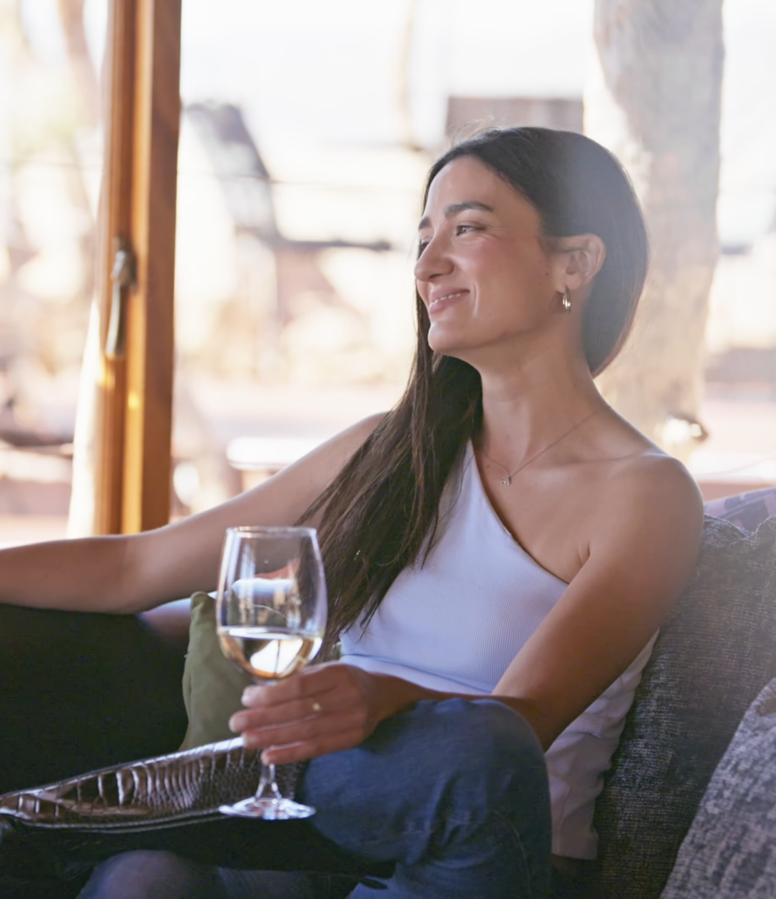 Woman relaxing on sofa holding a glass of white wine, smiling contentedly in a bright, airy lounge setting.