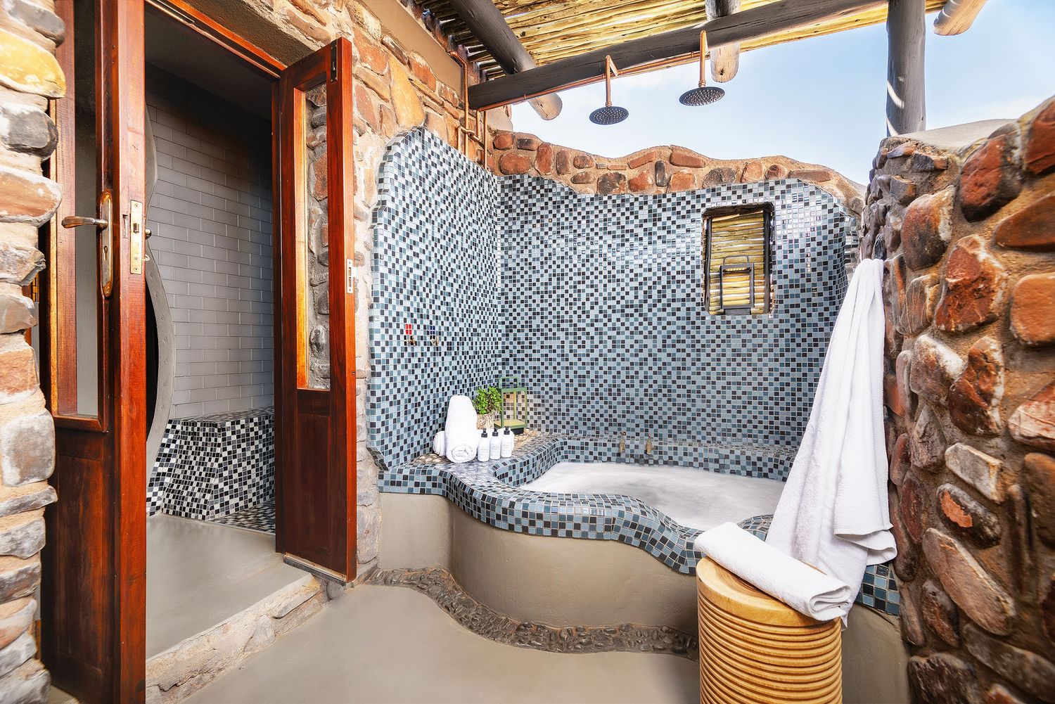 Outdoor safari bathroom with mosaic tiled bathtub, stone walls, and open-air shower area.