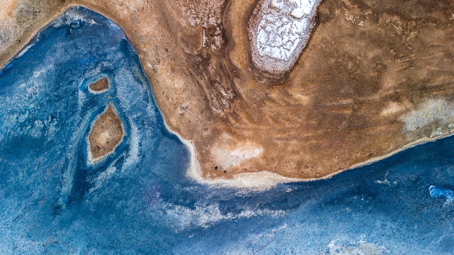 Aerial view of a stunning coastal landscape featuring vivid blue water meeting sandy brown earth with white salt deposits.