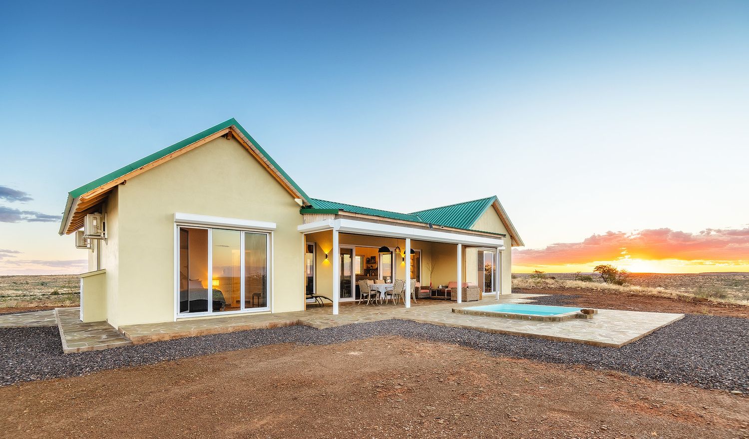 A yellow farmhouse with green roof and outdoor jacuzzi glowing under a vibrant sunset in an arid landscape.