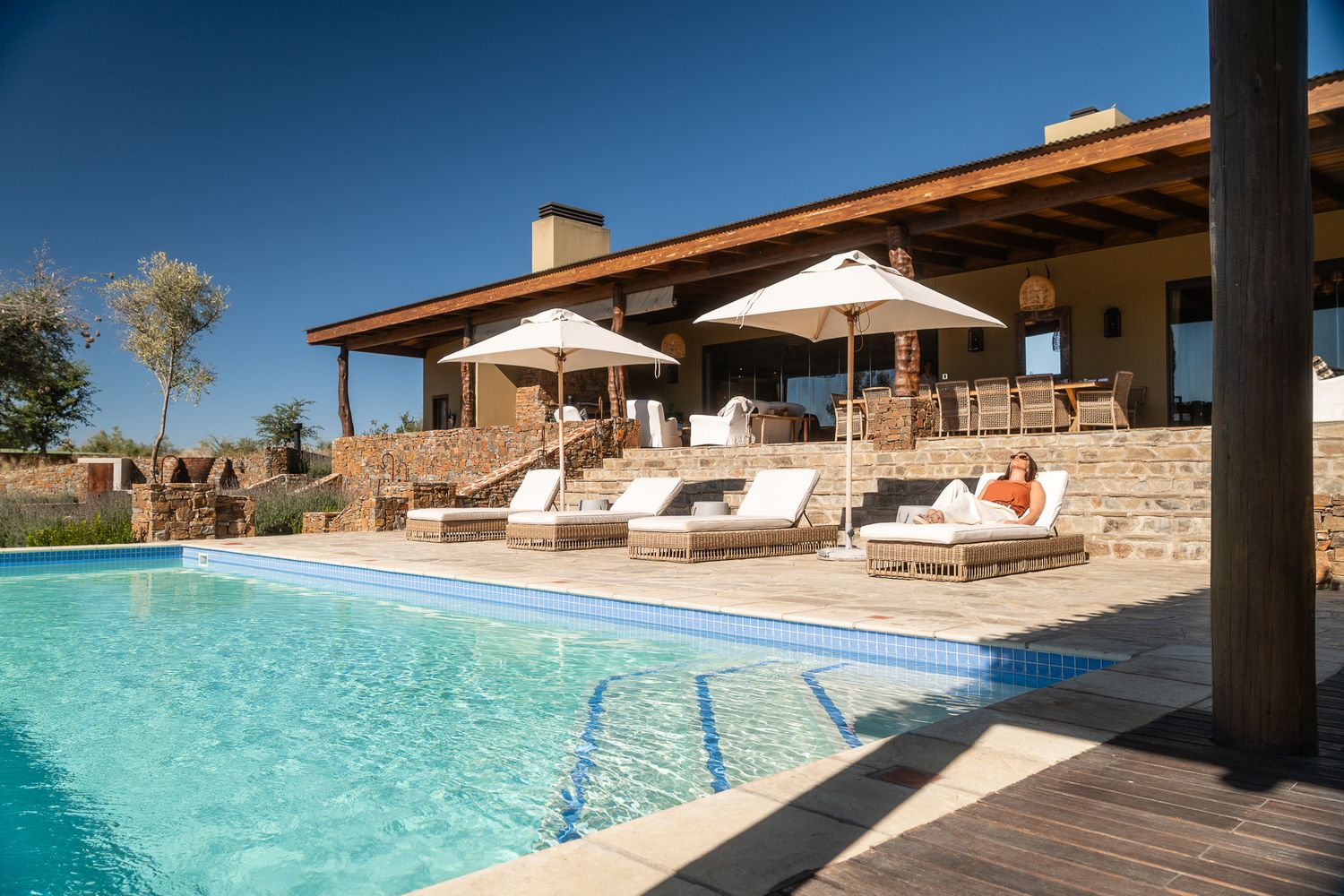 Luxury villa with sparkling pool, sun loungers, and umbrellas on a sunny terrace surrounded by stone walls.