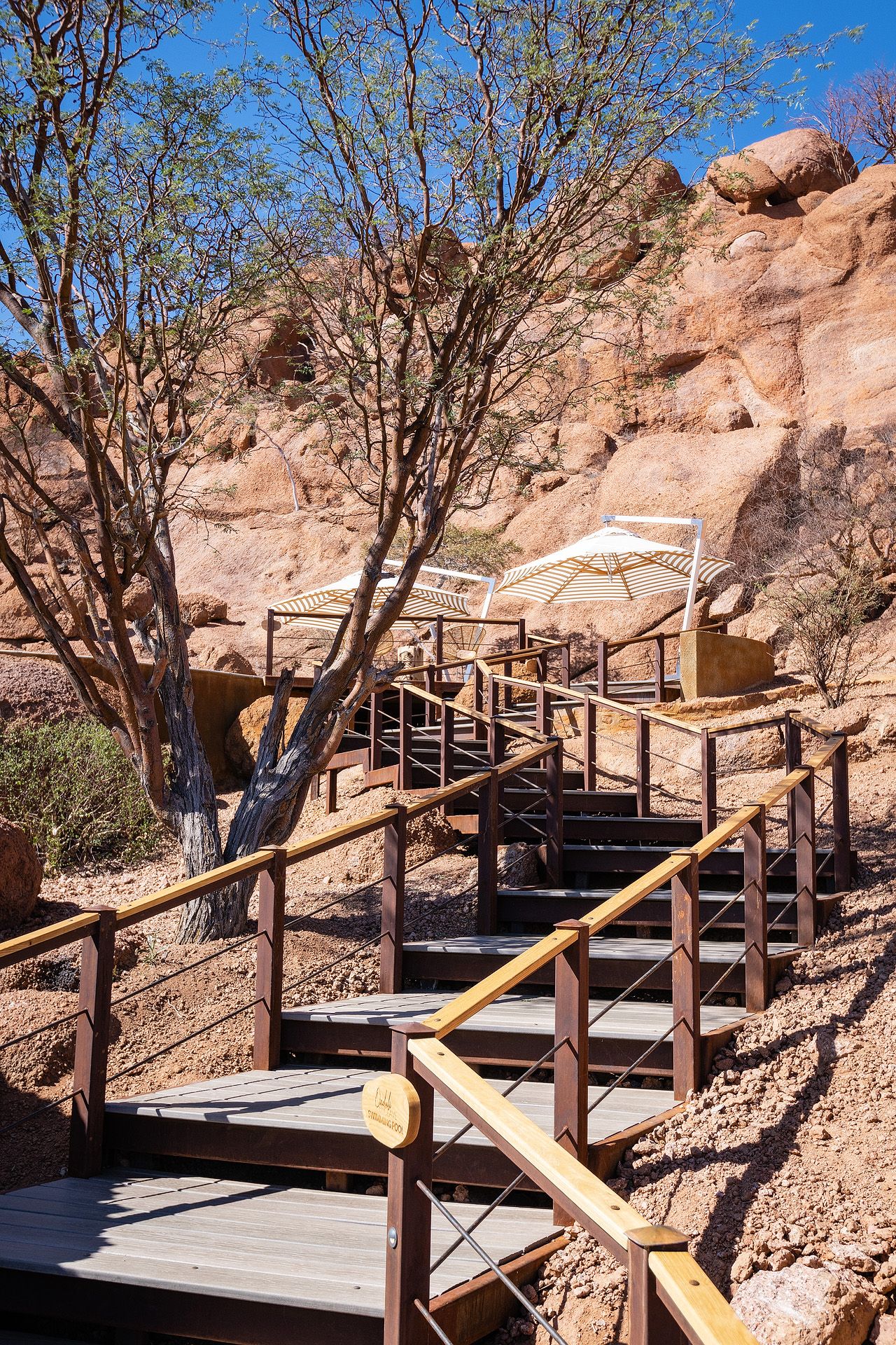 Wooden staircase with railings leading up rocky desert terrain with sparse trees and a white umbrella under blue sky.