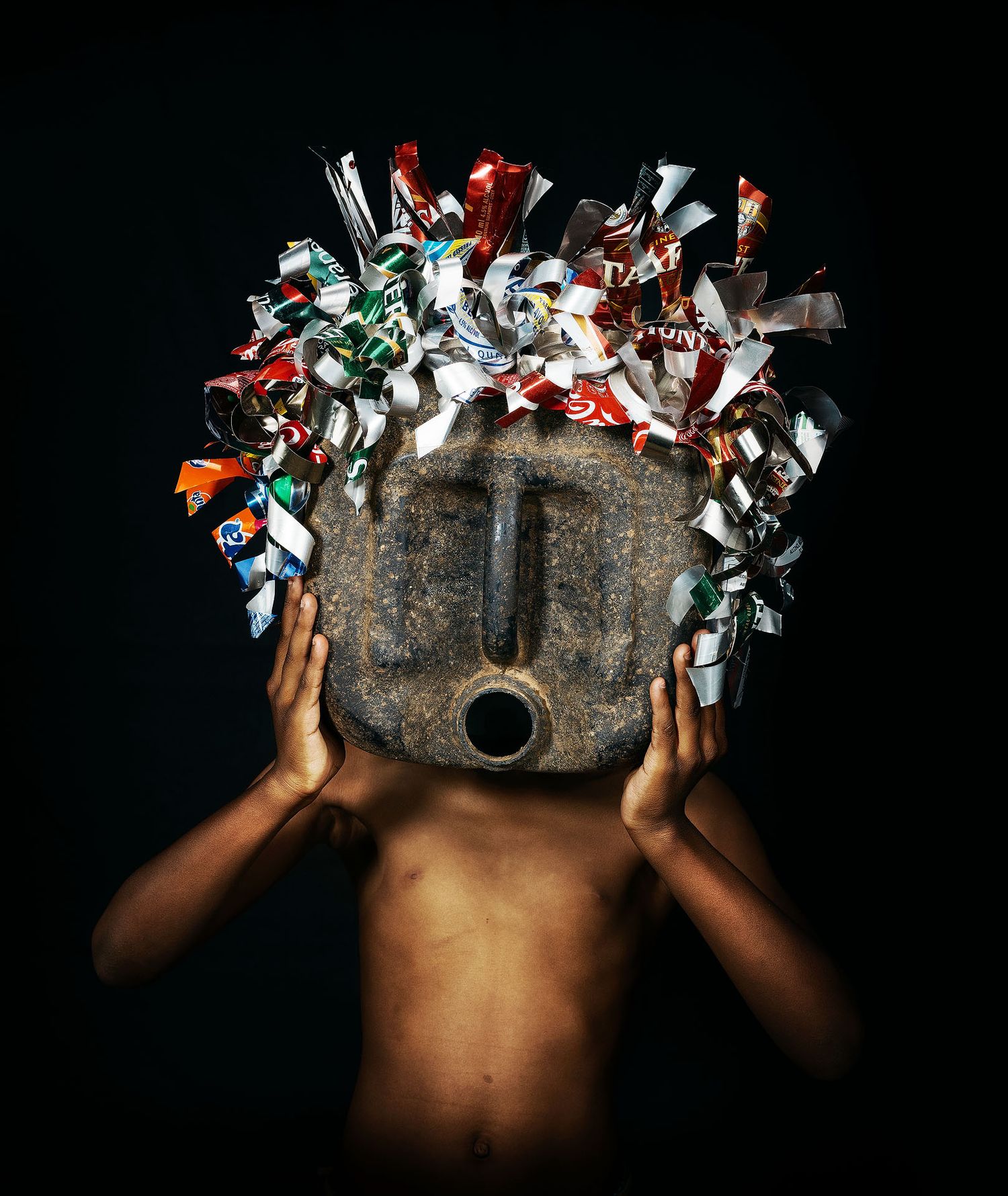 Person holding a tribal stone mask adorned with colorful candy wrappers against a dark background.