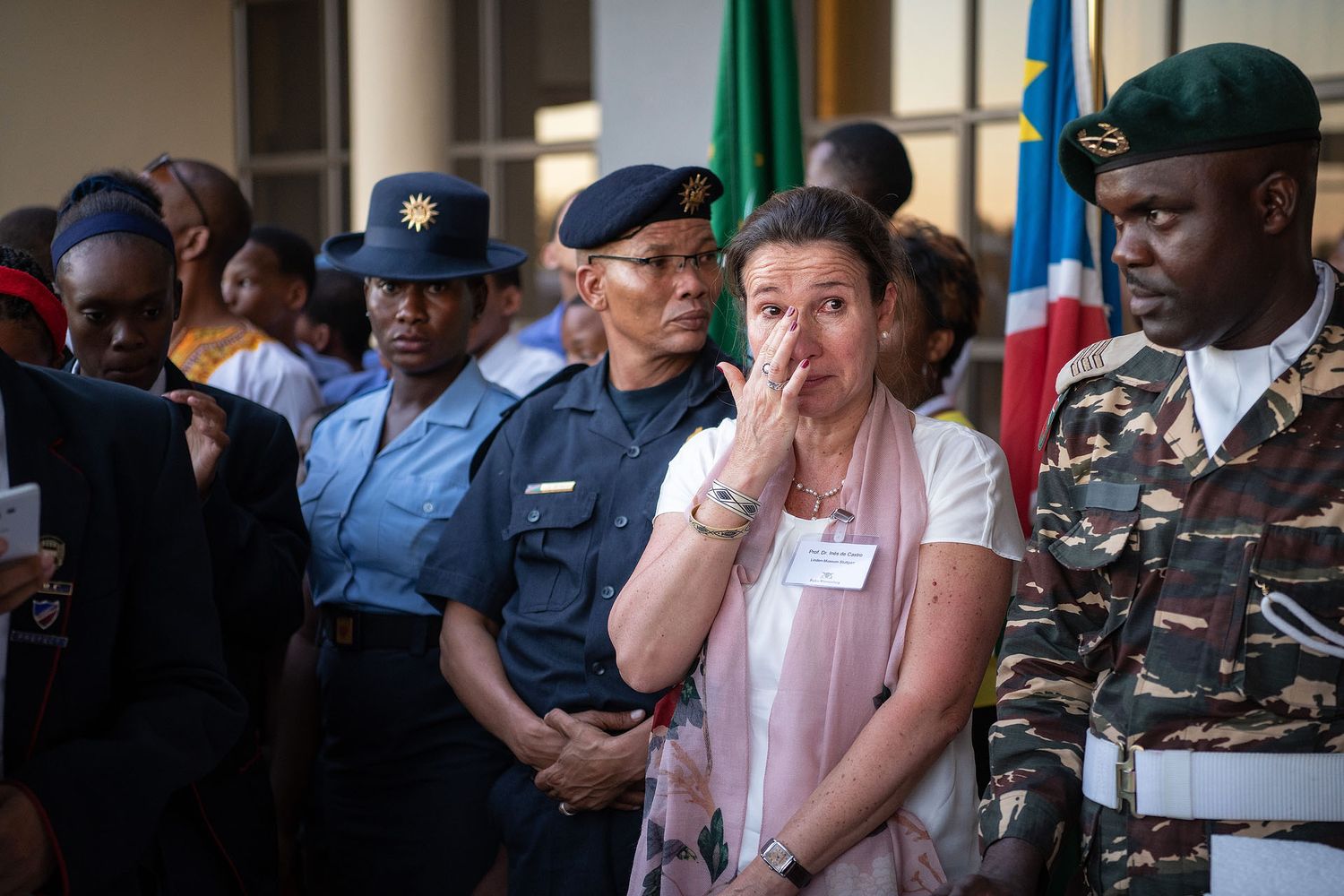 Emotional woman in pink blouse wipes tears at formal ceremony, flanked by military officers in uniform with flags.