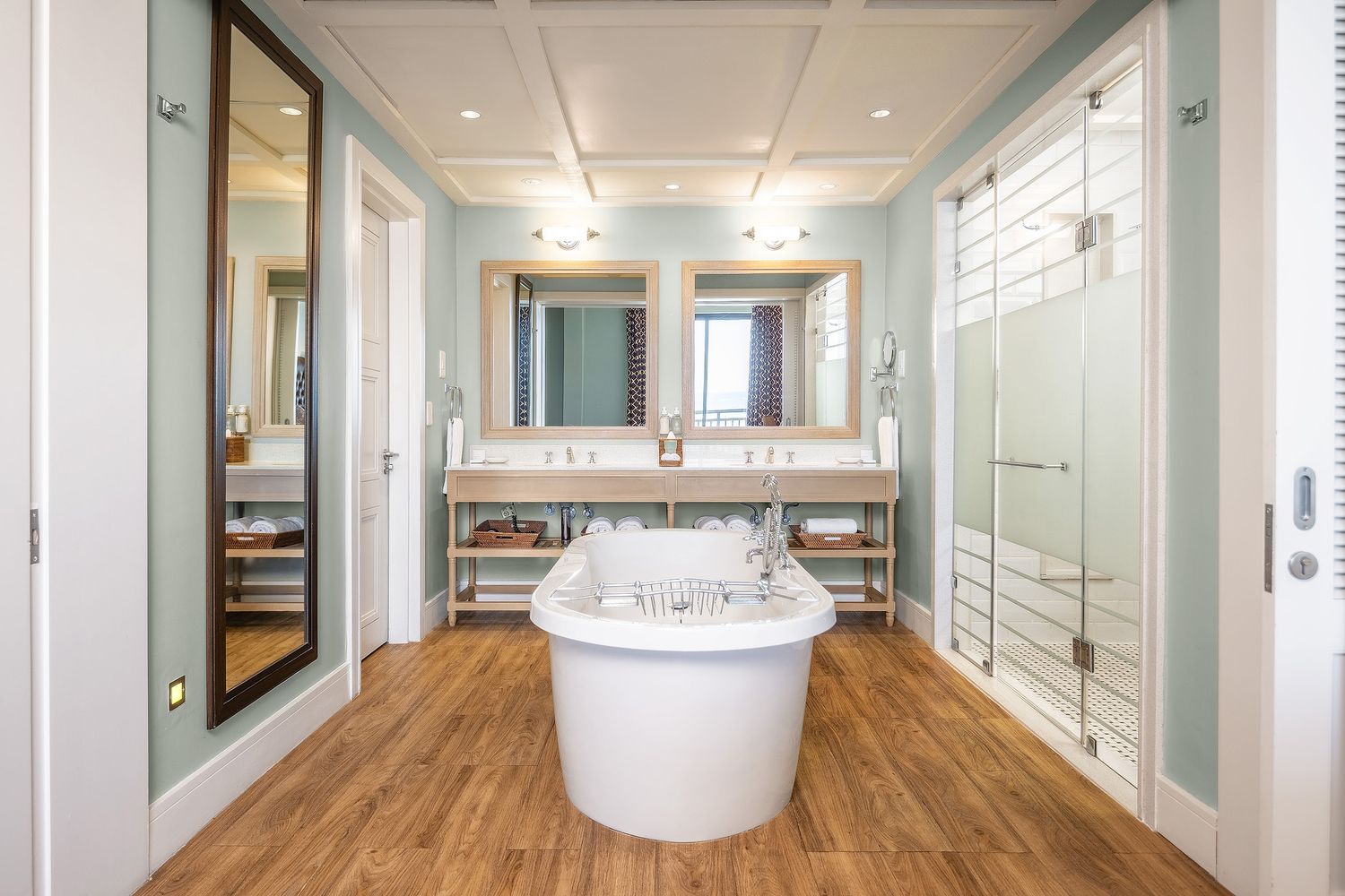 Elegant bathroom with freestanding soaking tub, dual wood-framed mirrors, glass shower, and warm hardwood floors.