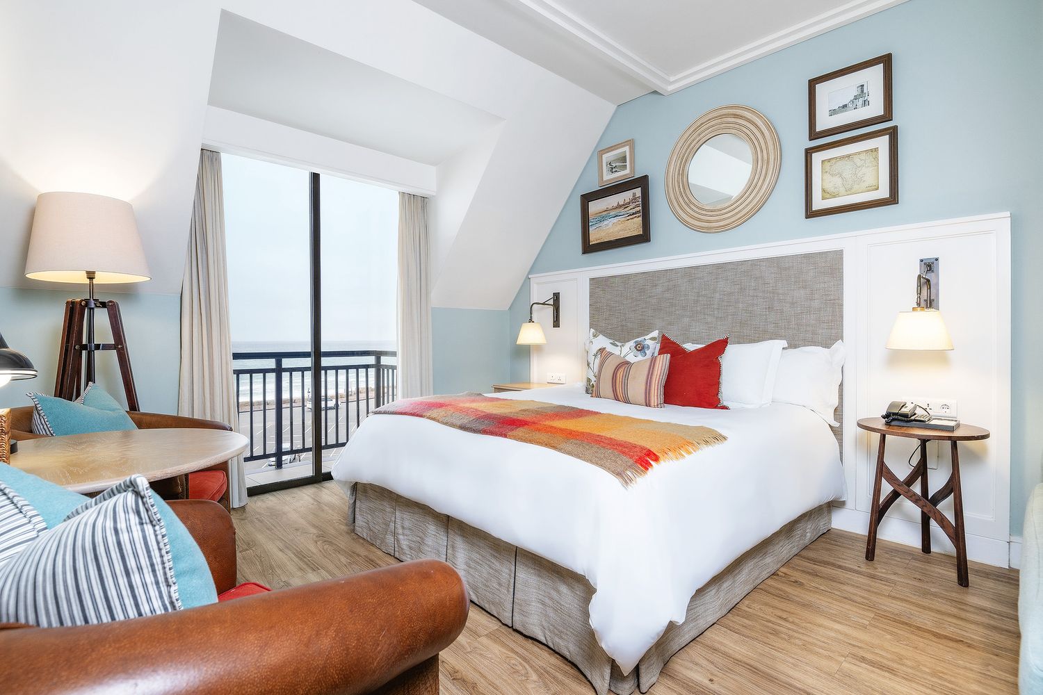 Bright hotel room with king bed, colorful throw, gray headboard, balcony view, hardwood floors, and coastal blue walls.