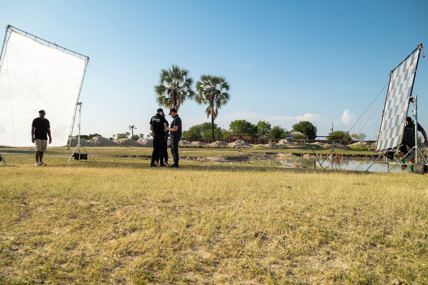 Film crew with reflectors on grassy field near palm trees and water on a sunny day.