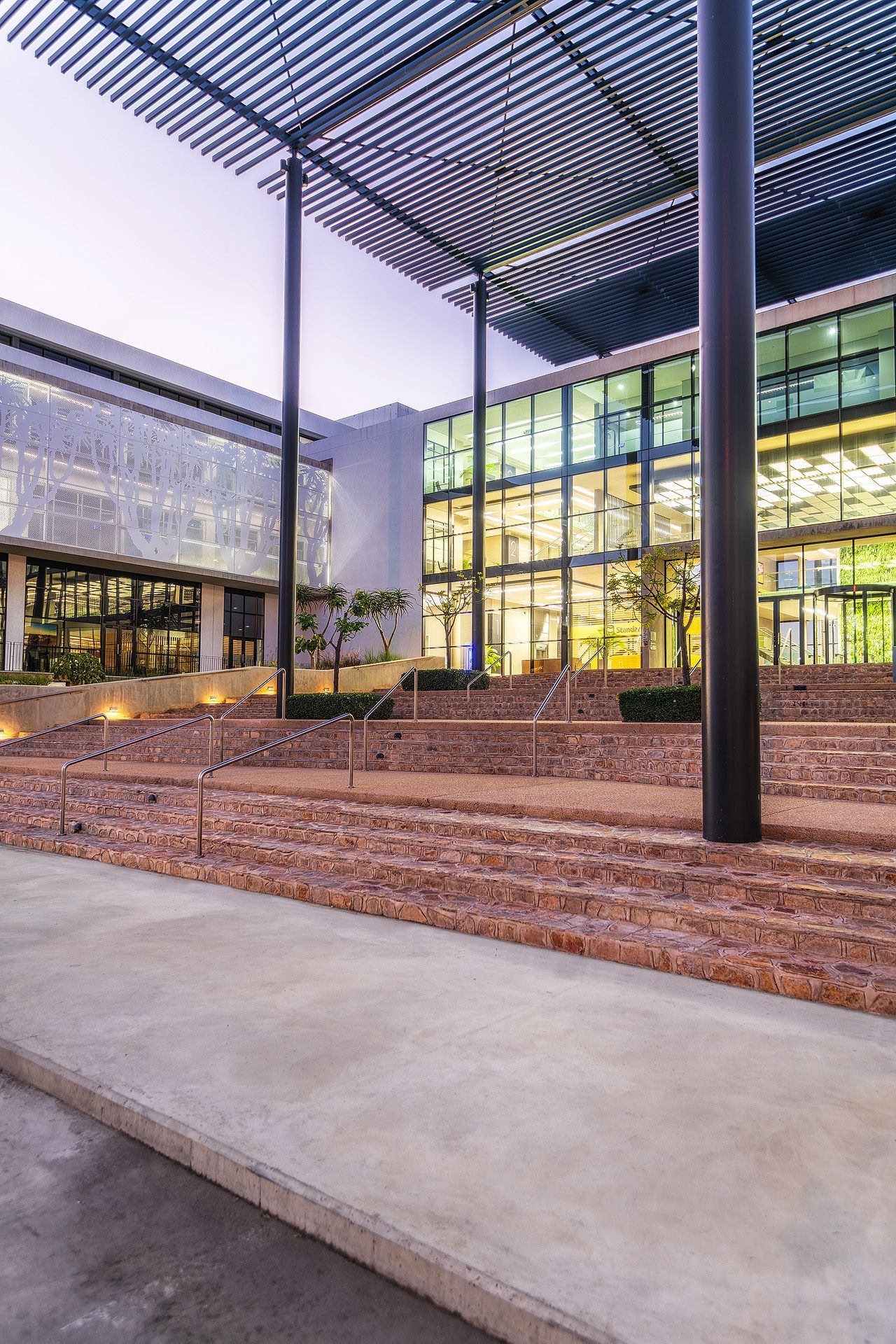 Modern building exterior with red brick stairs, glass facade, and pergola structure at dusk.