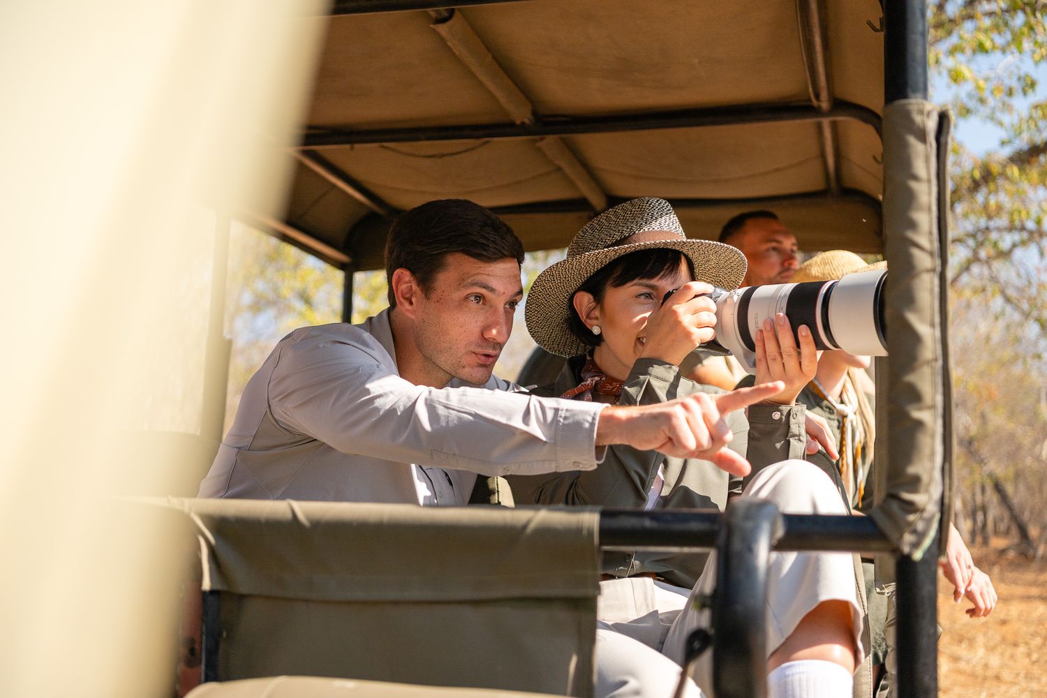 Tourists on a safari game drive, with a woman using a telephoto camera lens while a man points at wildlife.