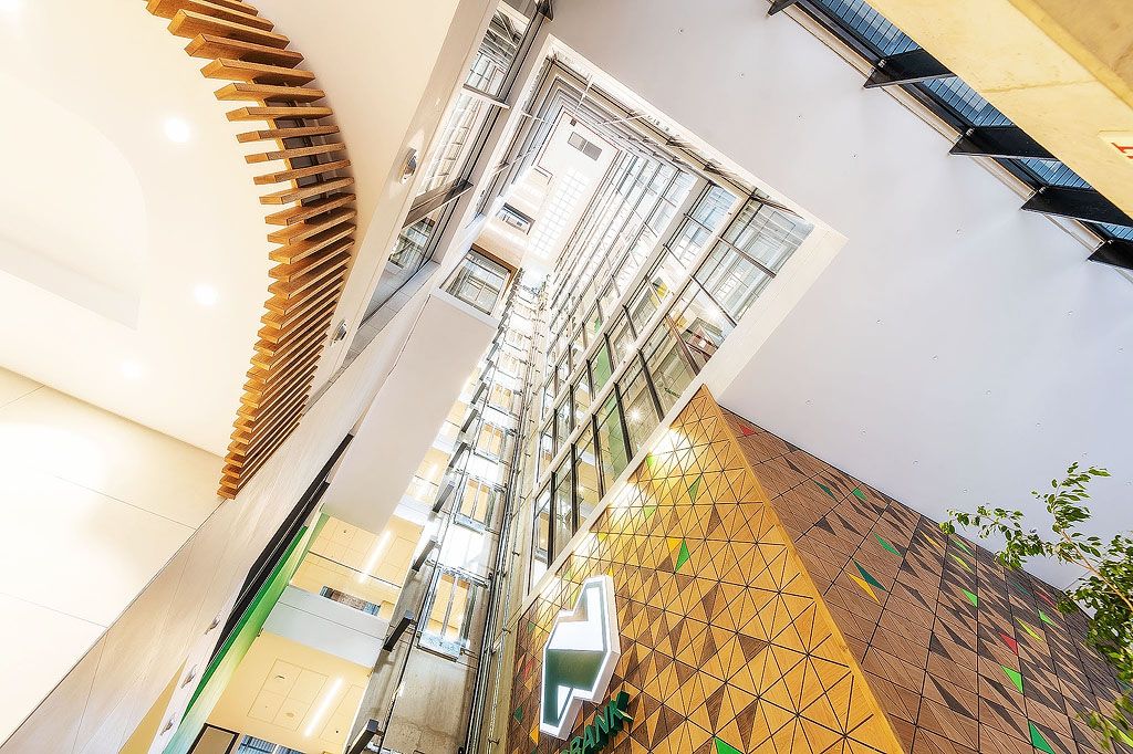 Dramatic upward view of modern office atrium with geometric wooden ceiling, glass elevators, and triangular wall tiles.