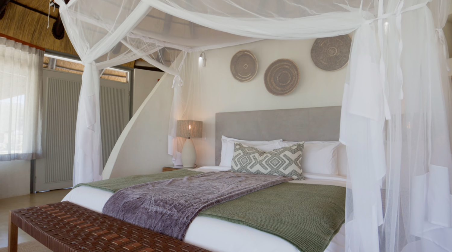 Elegant safari lodge bedroom with canopy bed, white mosquito net, green bedding, and decorative woven wall baskets.