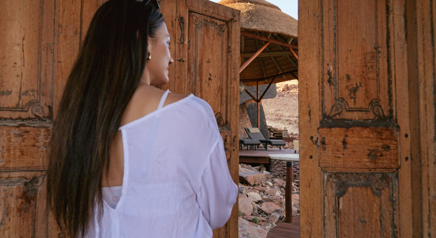 Woman in white off-shoulder top stands at rustic wooden doors overlooking a desert resort patio.