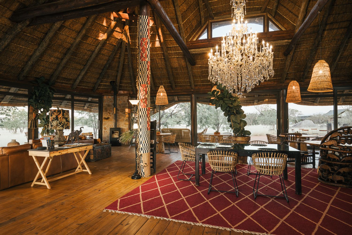 Elegant safari lodge interior with chandelier, red patterned rug, wooden floors, and open-air thatched roof overlooking nature.