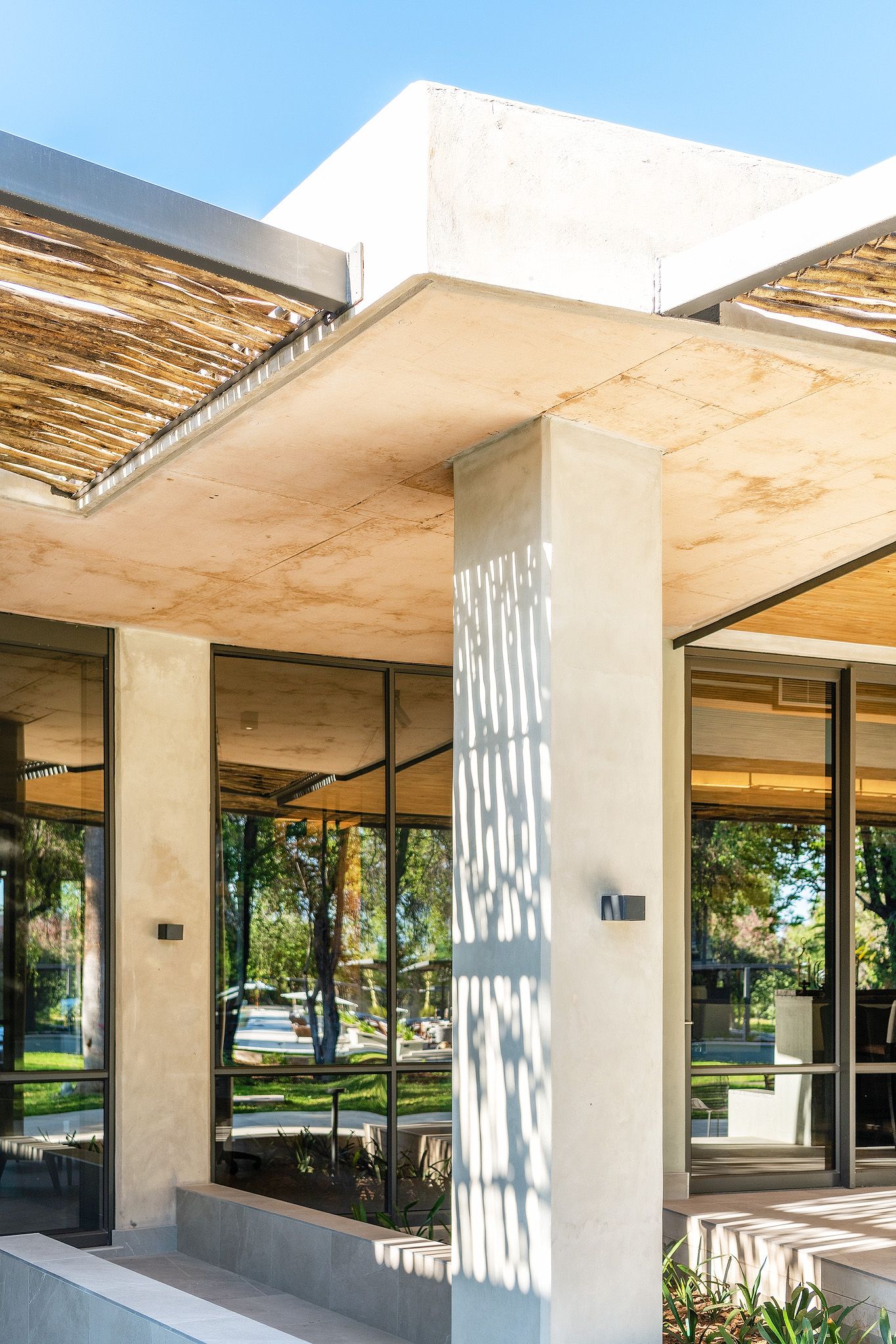 Modern building exterior with concrete pillars, glass walls, and a thatched pergola roof under a clear blue sky.