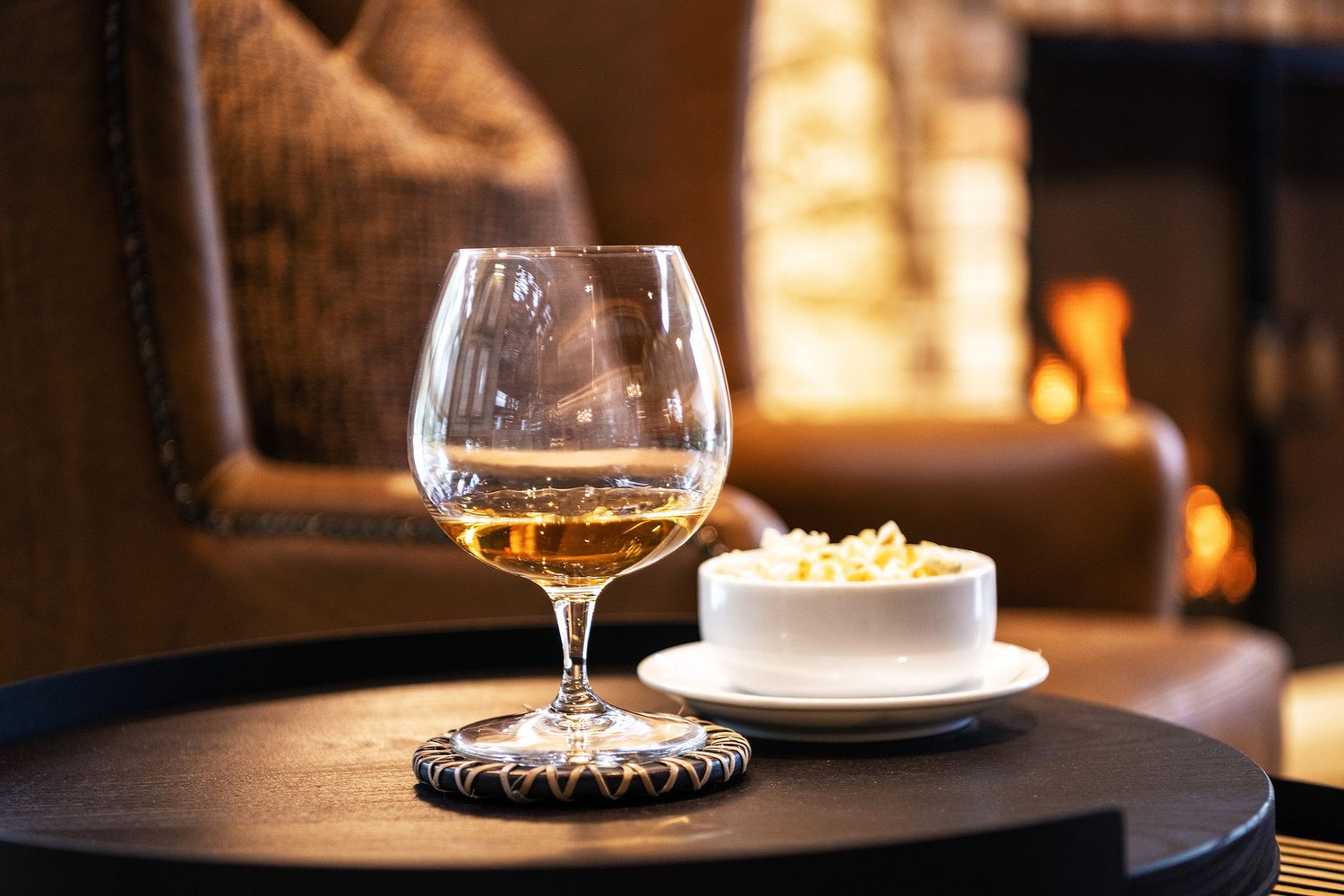 A glass of golden whisky beside a small bowl of snacks on a wooden table near a warm fireplace.