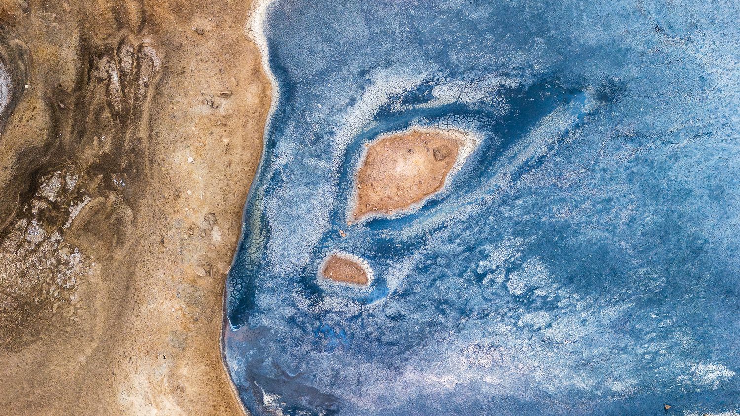 Aerial view of a stunning blue salt lake with brown sandy shores and oval island formations in arid landscape.