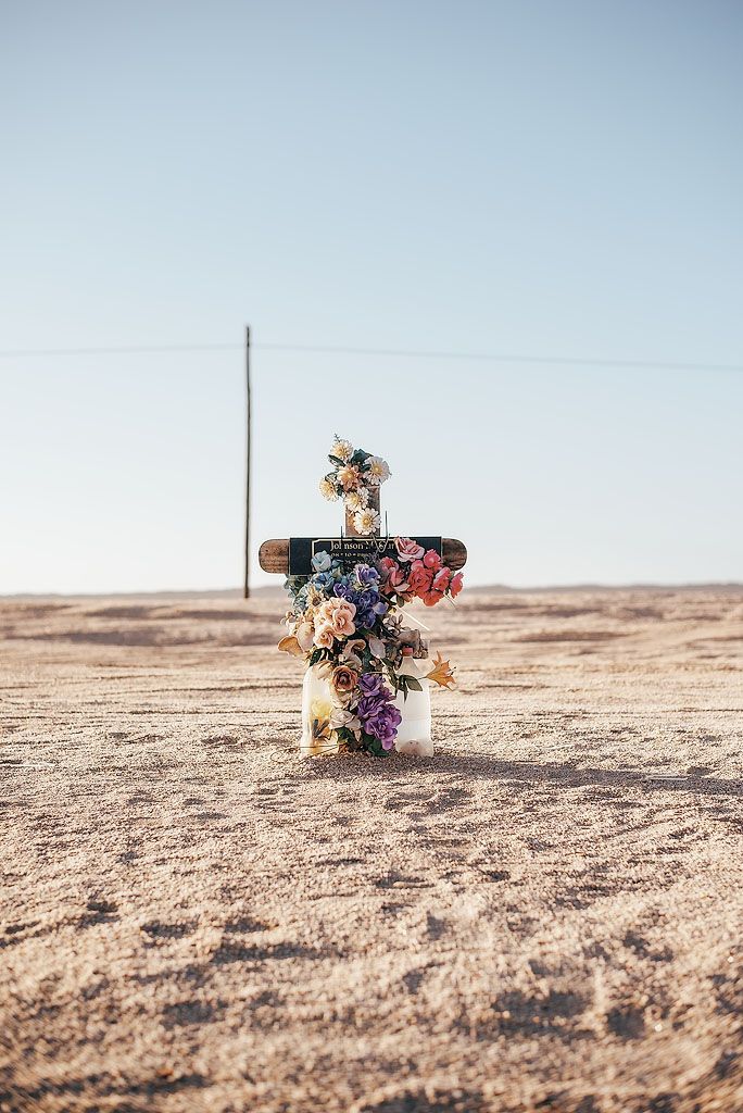 A flower-adorned roadside memorial cross stands alone in a vast, arid desert landscape under a clear sky.