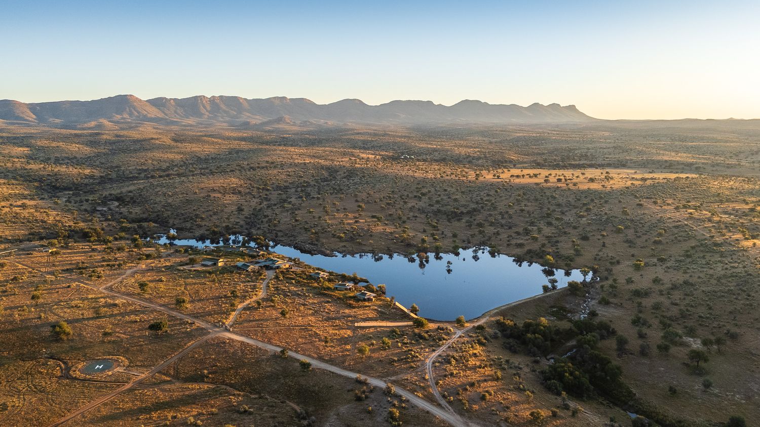 Aerial view of a serene lake surrounded by arid savanna landscape with mountain ranges under golden hour light.