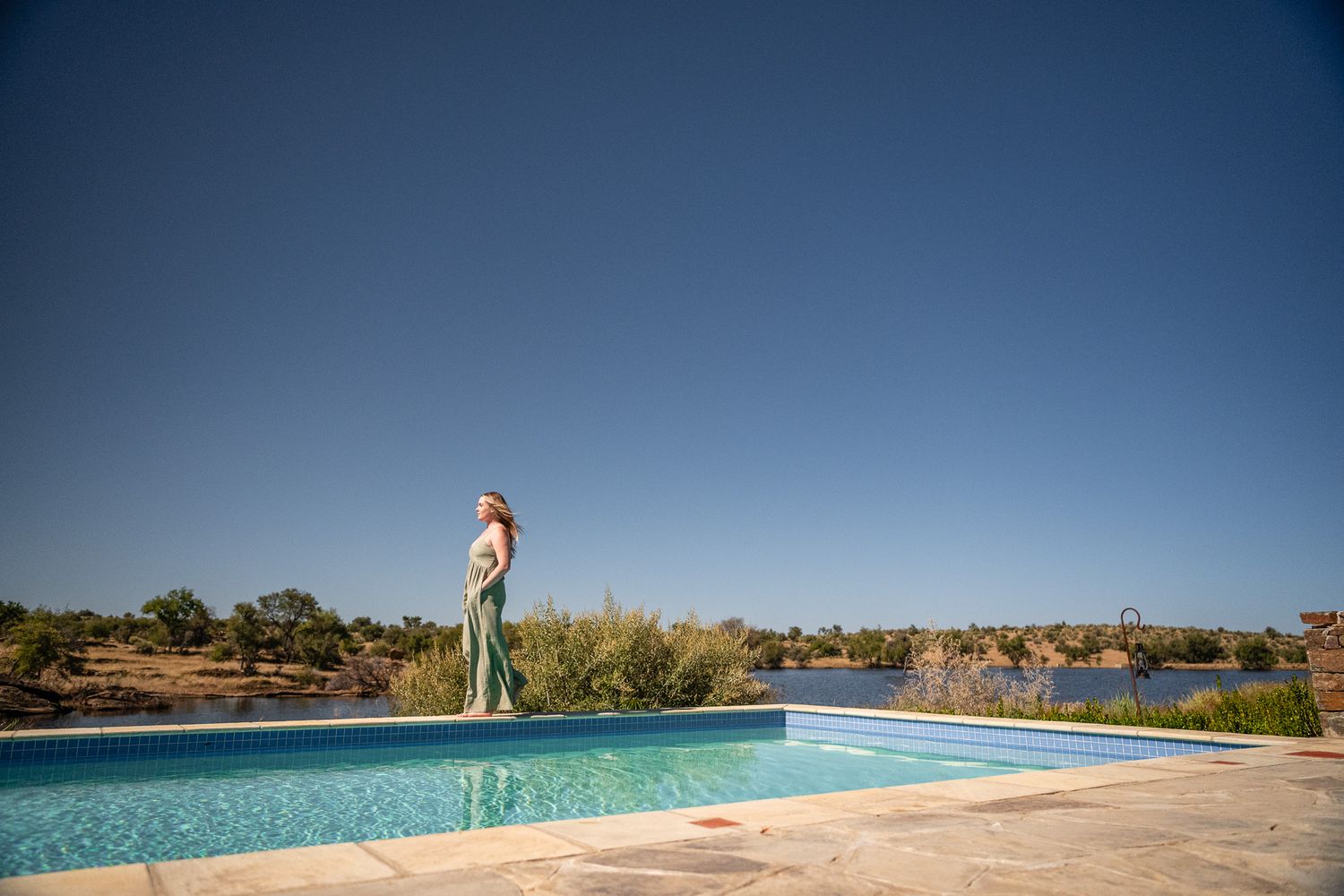 Woman in green dress standing beside a serene outdoor infinity pool under a clear blue sky in a sunny landscape.