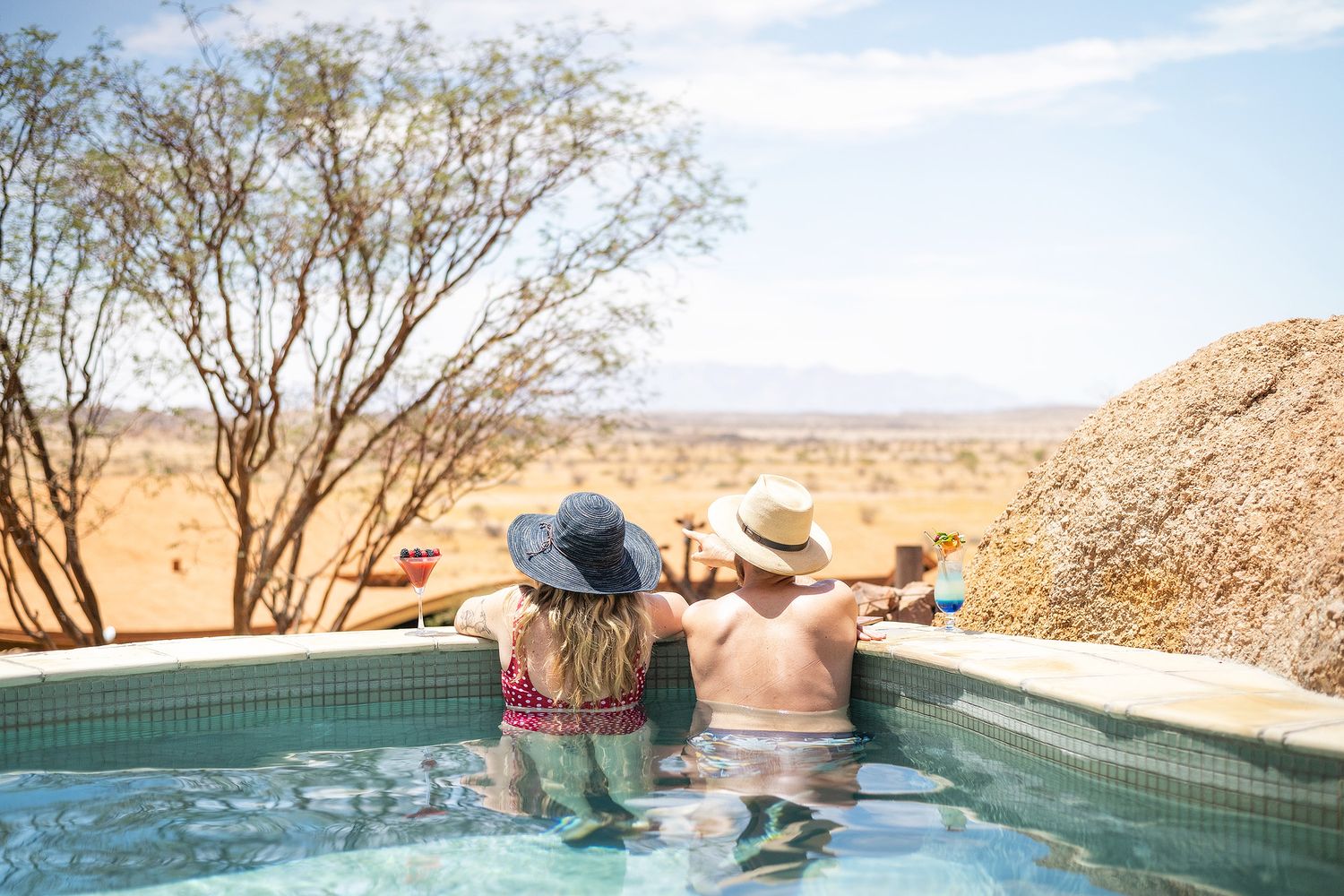 Couple relaxing in an outdoor pool with stunning desert landscape views, wearing sun hats on a sunny day.