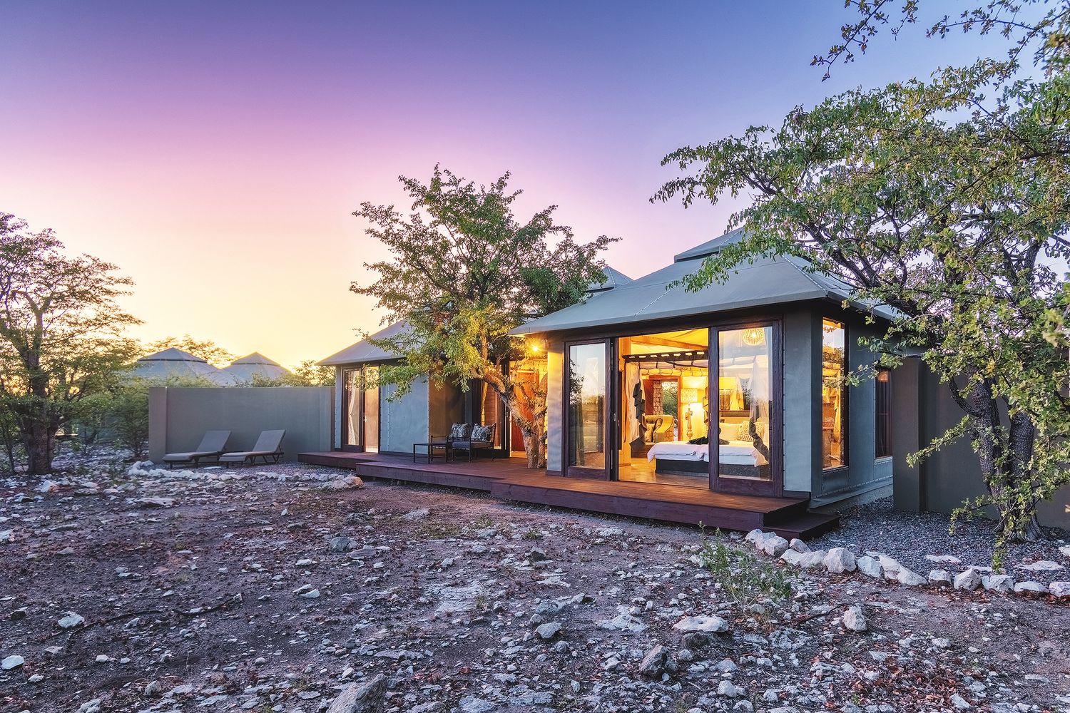 Modern safari lodge with glass walls and wooden deck glowing at dusk, surrounded by African bush under a purple sky.