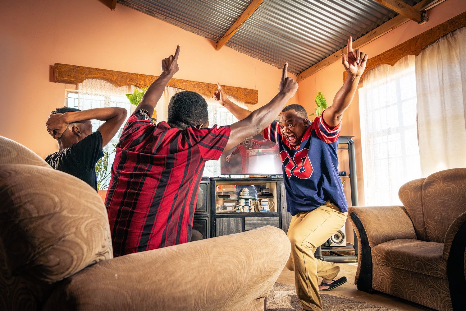 Three men celebrating with raised arms in a living room, wearing sports jerseys, expressing joy and excitement.