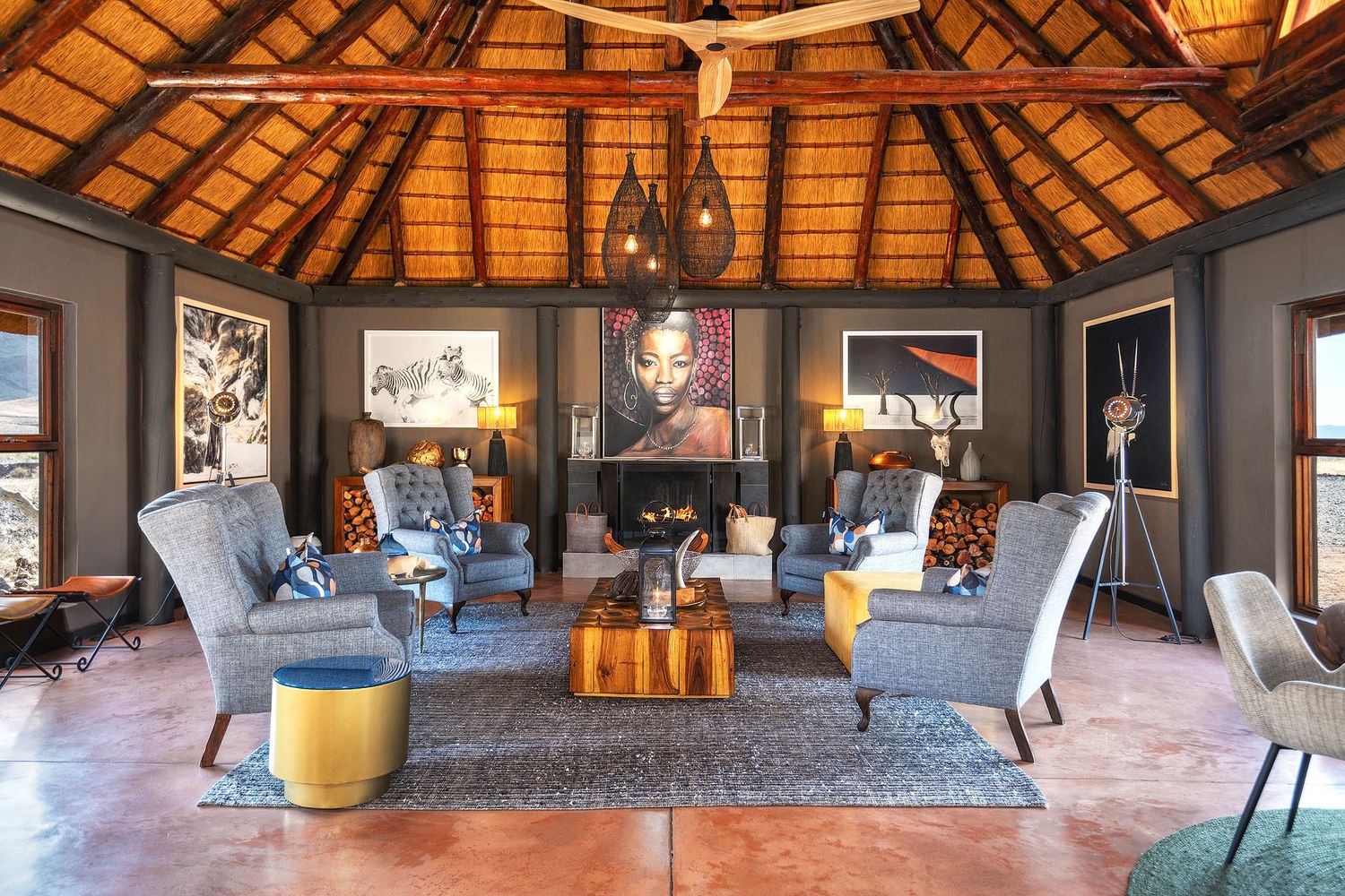 Stylish safari lounge with thatched roof, grey armchairs, wildlife artwork, and a lit fireplace.