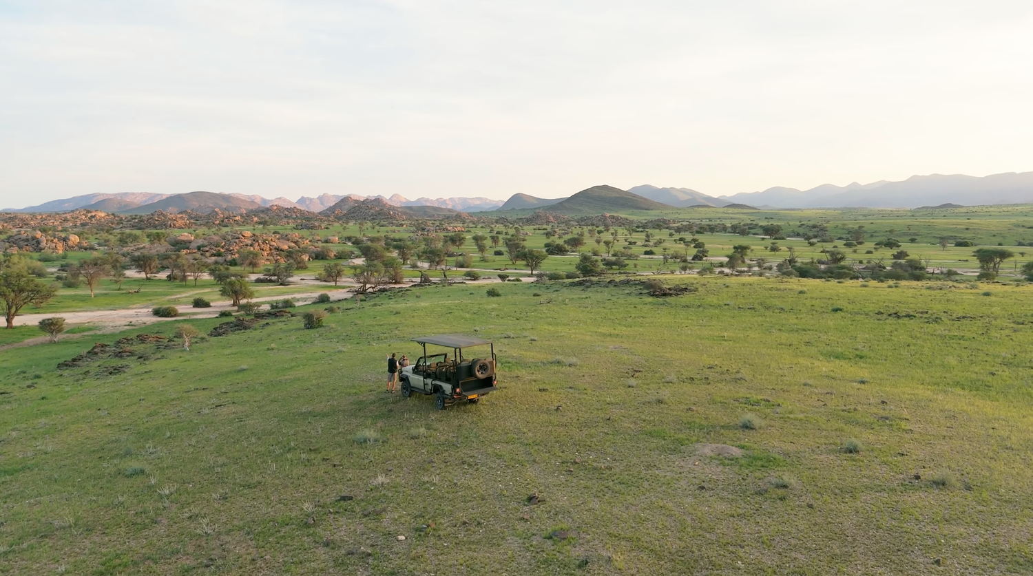 Aerial view of a safari camp surrounded by lush green savanna with scattered trees and rocky mountains in the background.