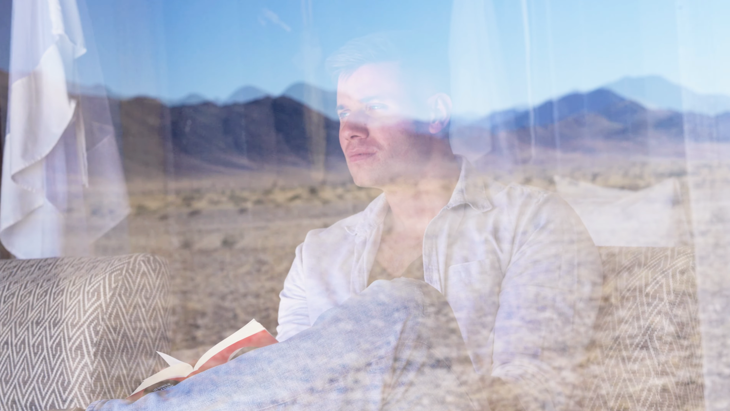Man in white robe reading a red book indoors with a desert mountain landscape visible through a window.