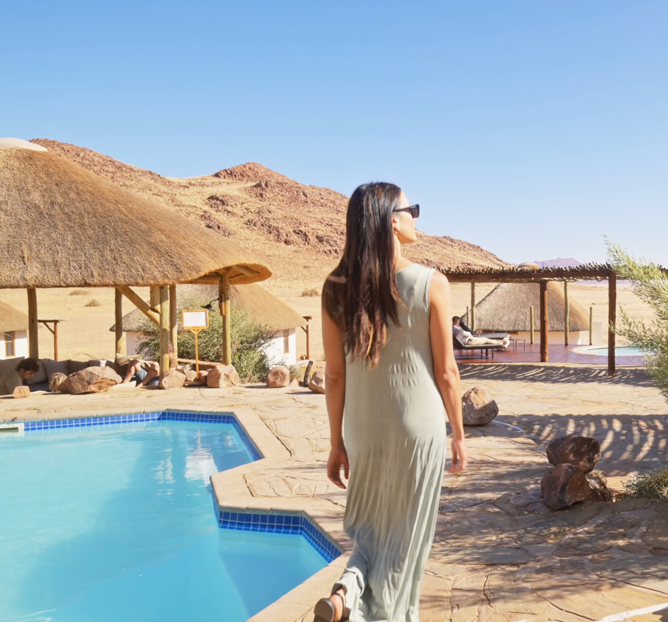 Woman in green dress stands by a desert lodge pool with thatched roofs and rocky hills under clear blue sky.