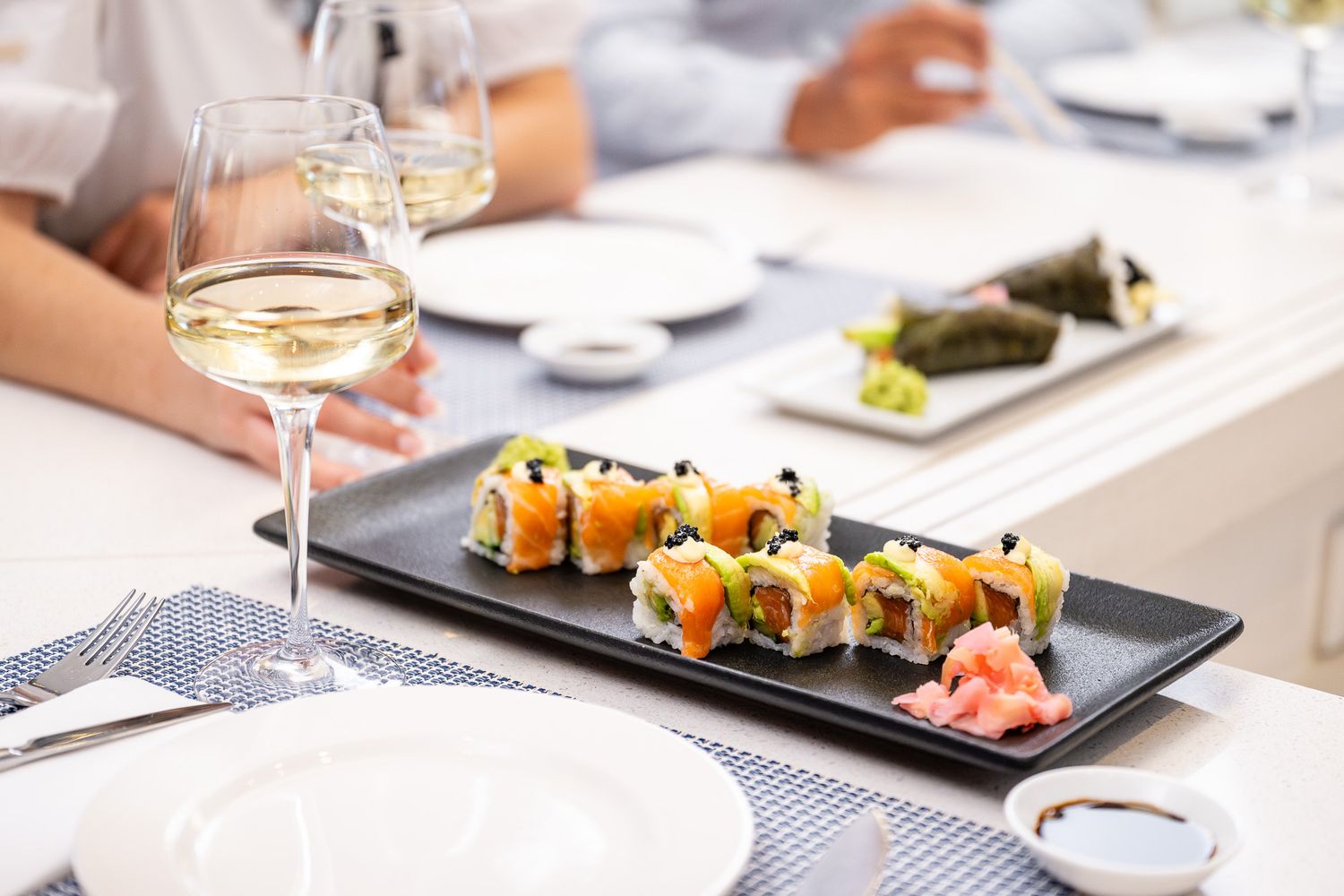 A plate of fresh salmon sushi rolls served with ginger and soy sauce, alongside a glass of white wine at a restaurant.